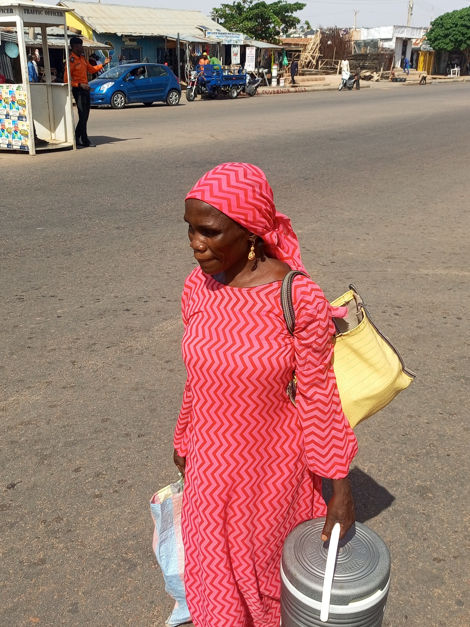 The primary subject of the image is an older woman walking on a paved road under bright sunlight. She is wearing a vibrant pink headscarf and a matching long-sleeved dress with a distinctive zigzag pattern. Gold-colored earrings adorn her ears. She carries a large, light yellow tote bag slung over her left shoulder, and in her hands, she holds a multi-colored woven bag and a large, cylindrical grey insulated container with a white handle. Her gaze is directed slightly downwards and to her left, indicating focus on her path.
In the mid-ground, a uniformed "TRAFFIC OFFICER" in an orange shirt and dark trousers stands beside a white booth, appearing to direct traffic or observe the street. A blue compact car, possibly a Toyota Yaris, is visible behind him. Further down the road, various roadside shops and structures are present, some appearing to be under construction with visible wooden scaffolding. A blue tricycle with a cargo bed and other motorcycles are also on the street. People are scattered throughout the background, engaged in various activities.
The setting appears to be a bustling street in an urban or semi-urban area, likely during the daytime given the harsh shadows and clear visibility. The weather is sunny. Visible text includes "TRAFFIC OFFICER" on the booth, "LIFE DAMY MEDICAL" on a sign, and faintly, "GOVERNOR 2023" spray-painted on the road, suggesting a political context. Posters featuring a man's face and the word "SALAH" are affixed to the traffic booth. The overall scene depicts daily life and commerce in a lively street environment.