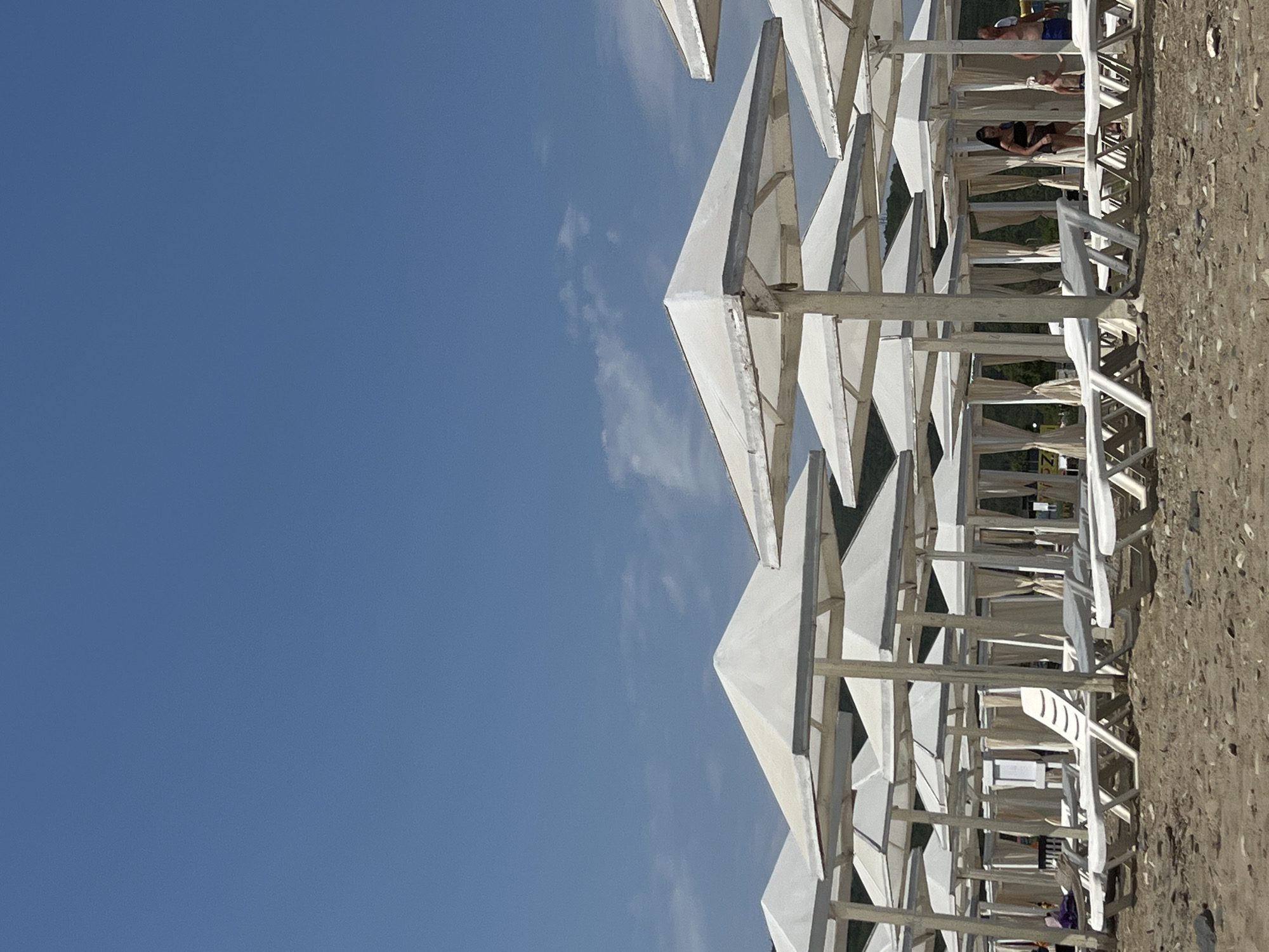The image captures a beach scene on a sunny day in Lermontovo, Russia. The foreground is dominated by rows of white beach umbrellas and empty lounge chairs on a gravelly beach. In the background, several cabanas with beige curtains are visible. A few people are present, including a woman in a black bikini and a man in blue shorts near the cabanas. The sky is a clear blue with a few scattered white clouds. The overall impression is one of a well-established resort beach, ready for visitors.