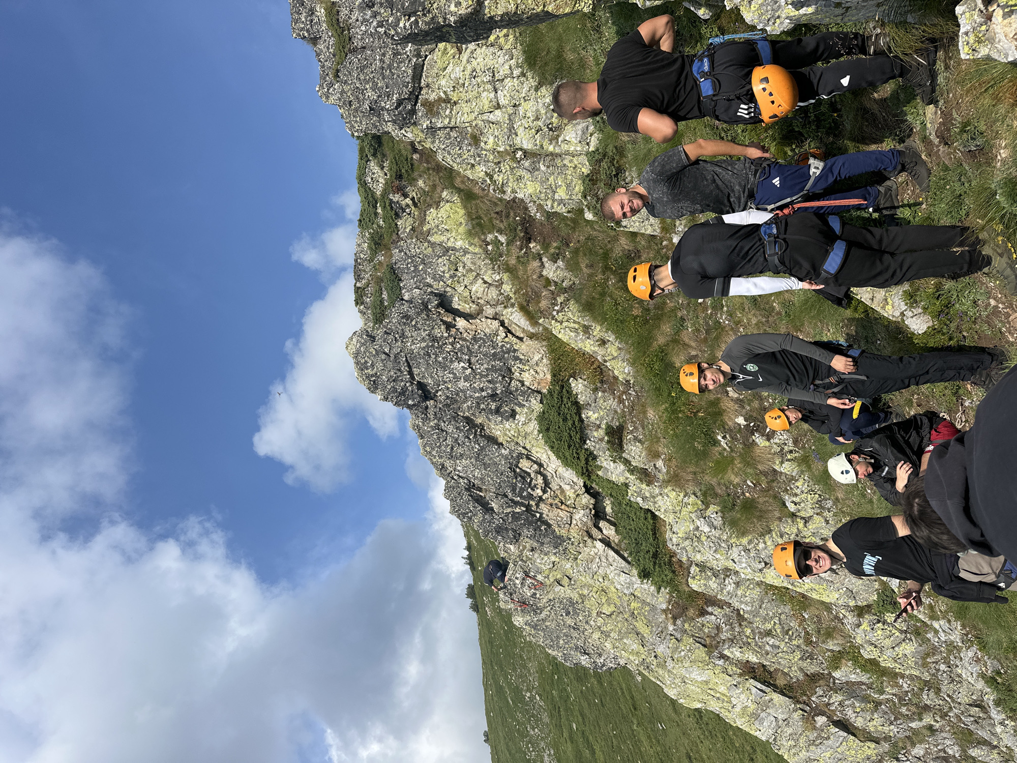 The image depicts a group of individuals at a rocky mountainous location. The individuals, all appearing to be male, are wearing climbing gear, including helmets and harnesses, suggesting they are engaged in rock climbing or a related activity. Some are standing, while others are seated. One individual is positioned higher up on a rock face, seemingly climbing. The environment is characterized by steep, rocky slopes covered in sparse vegetation, with a clear blue sky and scattered clouds overhead. The terrain suggests a natural, outdoor setting, possibly a hiking or climbing trail. The individuals are equipped with backpacks and ropes, further indicating preparation for an outdoor adventure. The overall scene captures a moment of activity and preparation in a rugged natural environment.