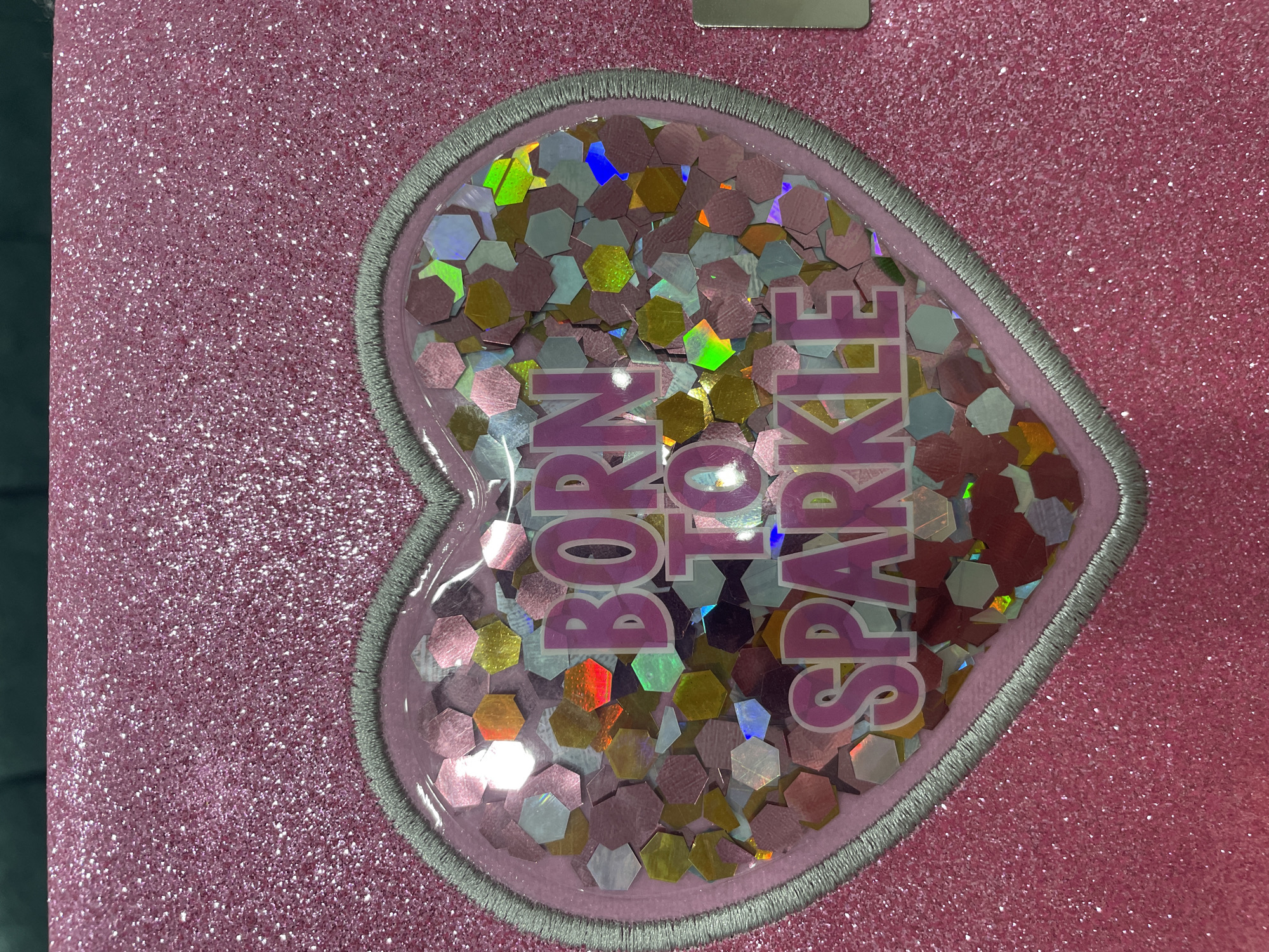 The image shows a close-up of a pink glittery surface with a heart-shaped appliqué. Inside the heart is a clear plastic window filled with hexagonal sequins in various colors, including pink, gold, silver, and iridescent hues. The words "BORN TO SPARKLE" are printed in pink lettering on the sequins. The background is a dark green surface. The image is a product shot and does not depict any specific location in Thuận An, Vietnam.