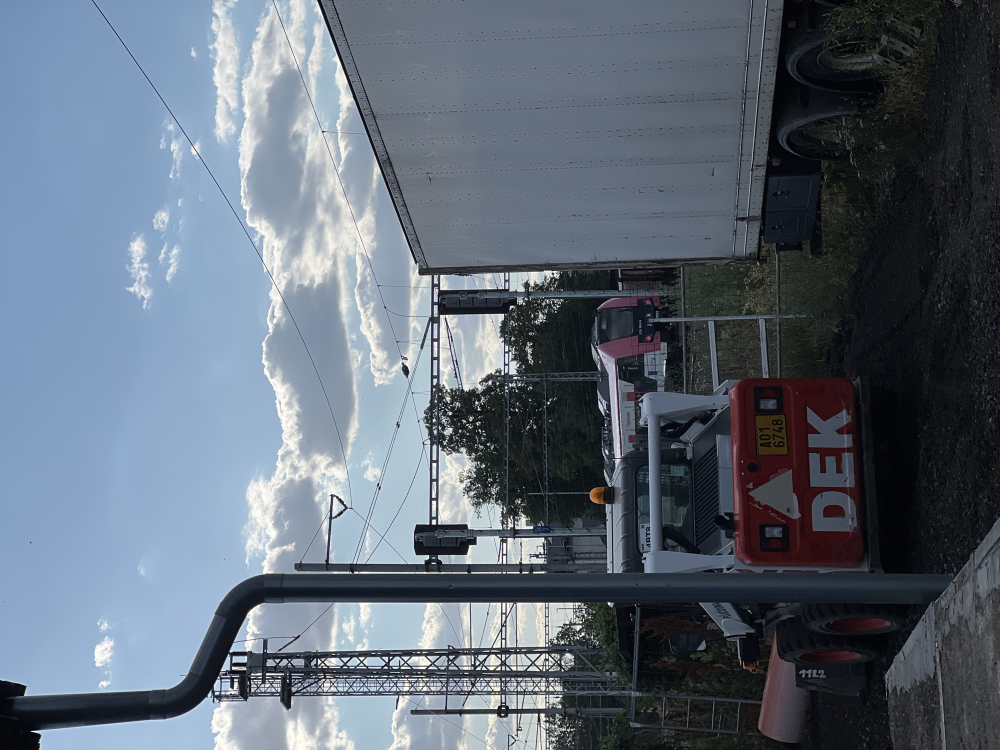 The image shows a construction site in Újezd u Brna, Czechia. In the foreground, a small orange and white skid steer loader with the text "DEK" on its rear is parked on dark soil. The loader has its engine running, indicated by a flashing orange light on top. Behind the loader, a white semi-trailer truck is parked. In the background, a red and white train is visible on a track, alongside overhead power lines and a lattice tower. The sky is blue with scattered white clouds, and there are trees on the horizon. The overall scene suggests ongoing construction or maintenance work near a railway line.