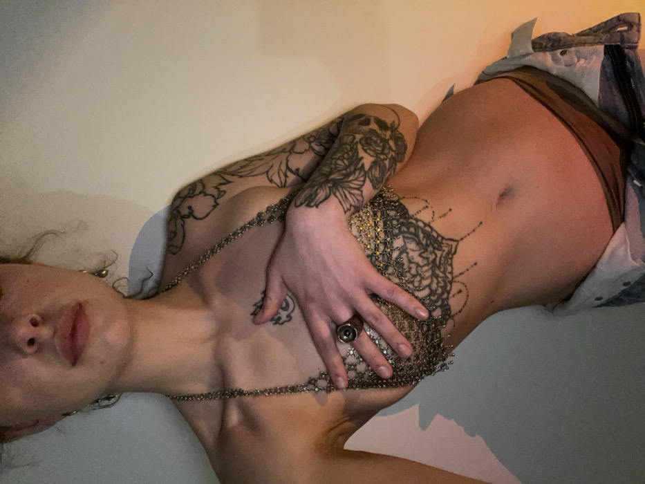 A woman lies on her side against a light-colored wall. She has extensive tattoos on her arm and chest, and a mandala-style tattoo on her stomach. She is partially clad in what appears to be a metal chainmail-like garment that covers her chest and midriff. A large ring is visible on her hand, which rests on the garment. Her face is in profile, showing red lips and a nostril piercing. Denim clothing is visible at her hip.