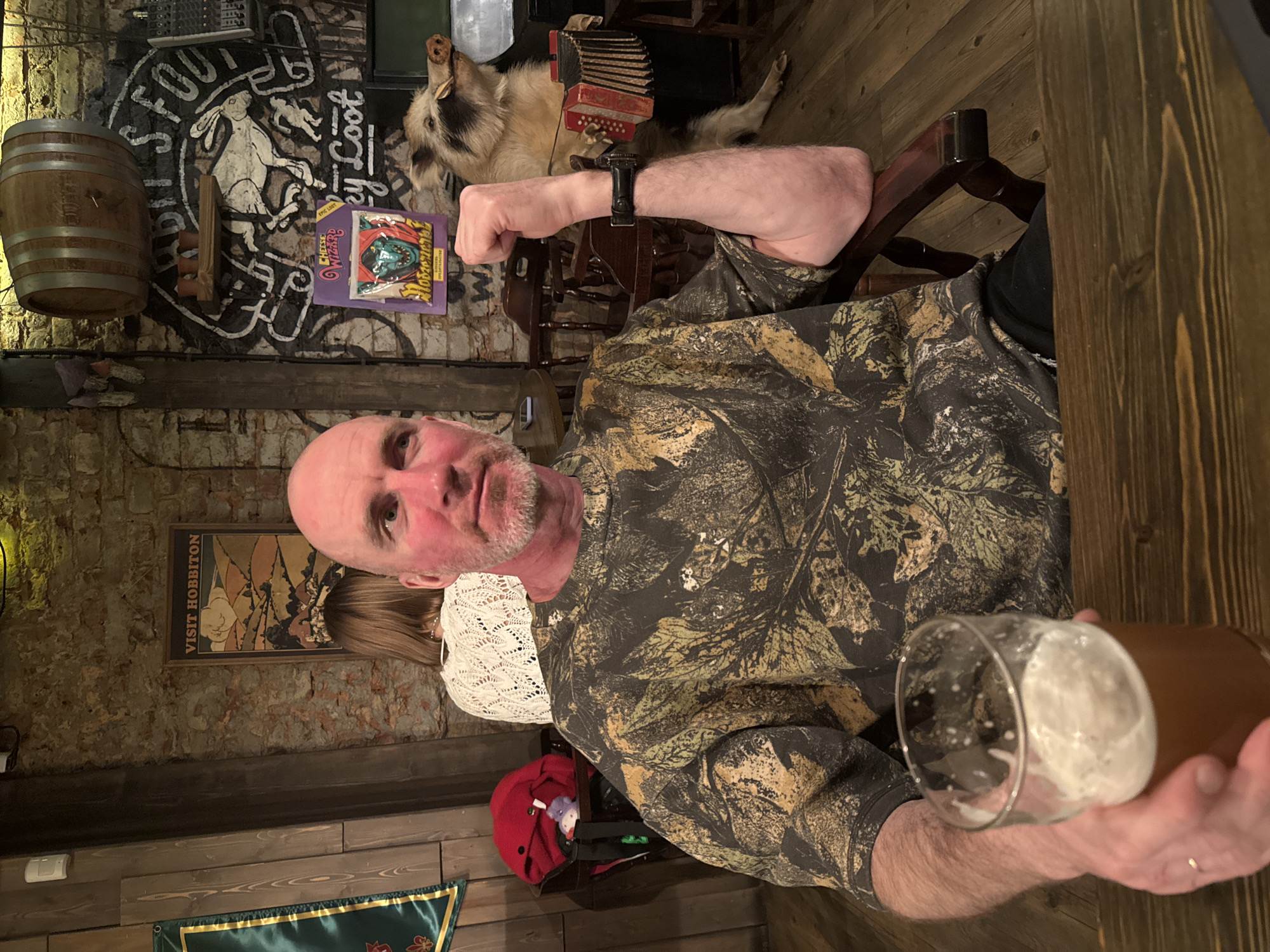 The image shows a man sitting at a wooden table, holding a pint of beer. He is wearing a camo-patterned shirt and a black wristwatch. The background includes a wall with exposed brick and wooden paneling. There is a barrel and an animal taxidermy with an accordion on the wall. A poster that reads "Visit Hobbiton" is present. The image was taken in Saint Petersburg, Russia, specifically at The Red Button Bar (Krasnaya Knopka Bar).