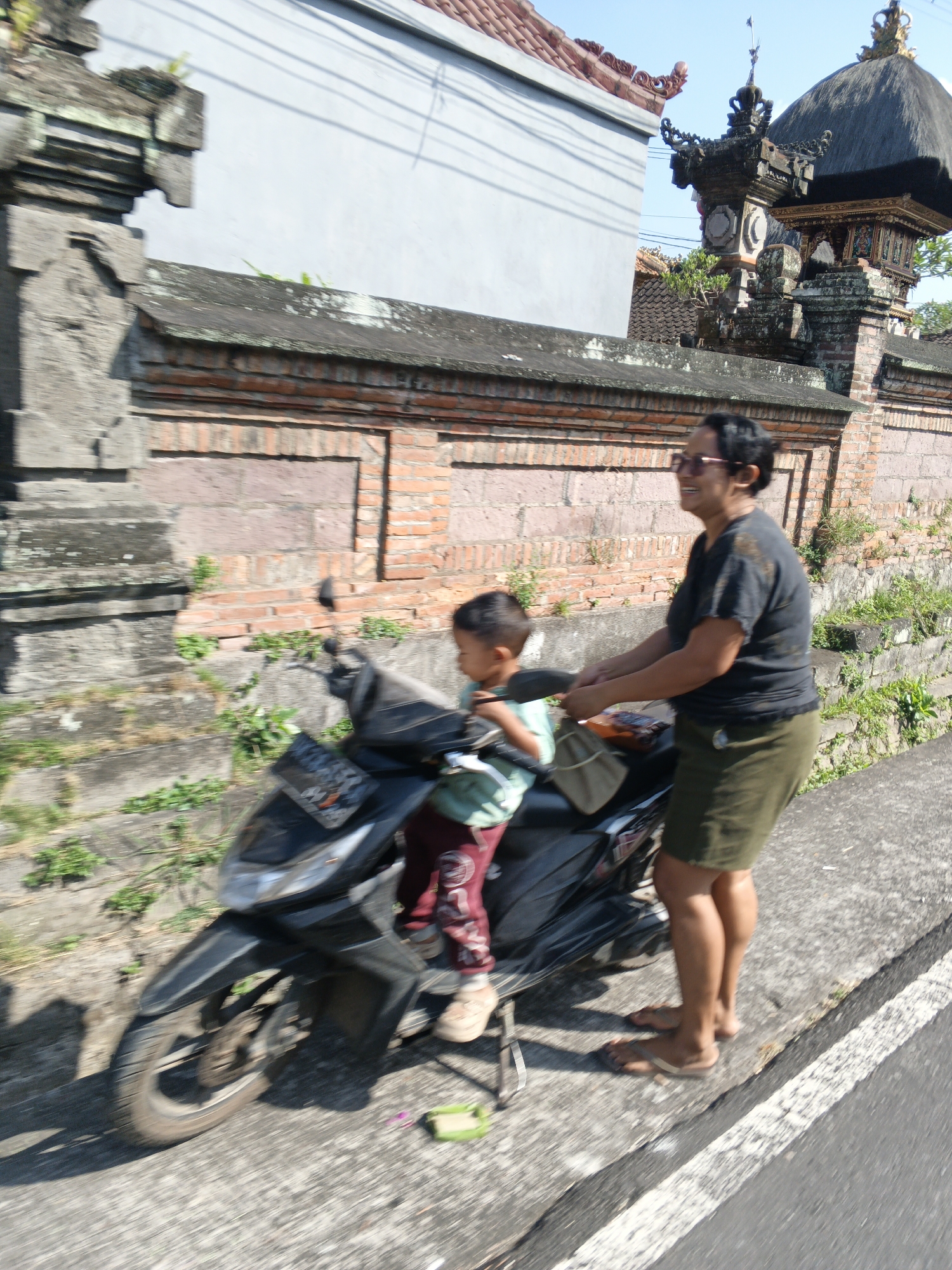 In Abiansemal, Indonesia, a woman is riding a motorcycle with a young child seated in front of her. The woman, wearing a dark t-shirt and a green skirt, is looking forward and smiling. She has dark hair and is wearing glasses. The child, dressed in a light green shirt and red patterned pants, is seated in front of her. The motorcycle is black and is parked on the side of a road next to a brick wall. The wall has a stone pillar on the left and an ornate structure with a thatched roof on the right. Green vegetation is growing along the base of the wall. The road surface is visible in the foreground and on the right side of the image. The scene appears to be outdoors during the day, under a clear sky.