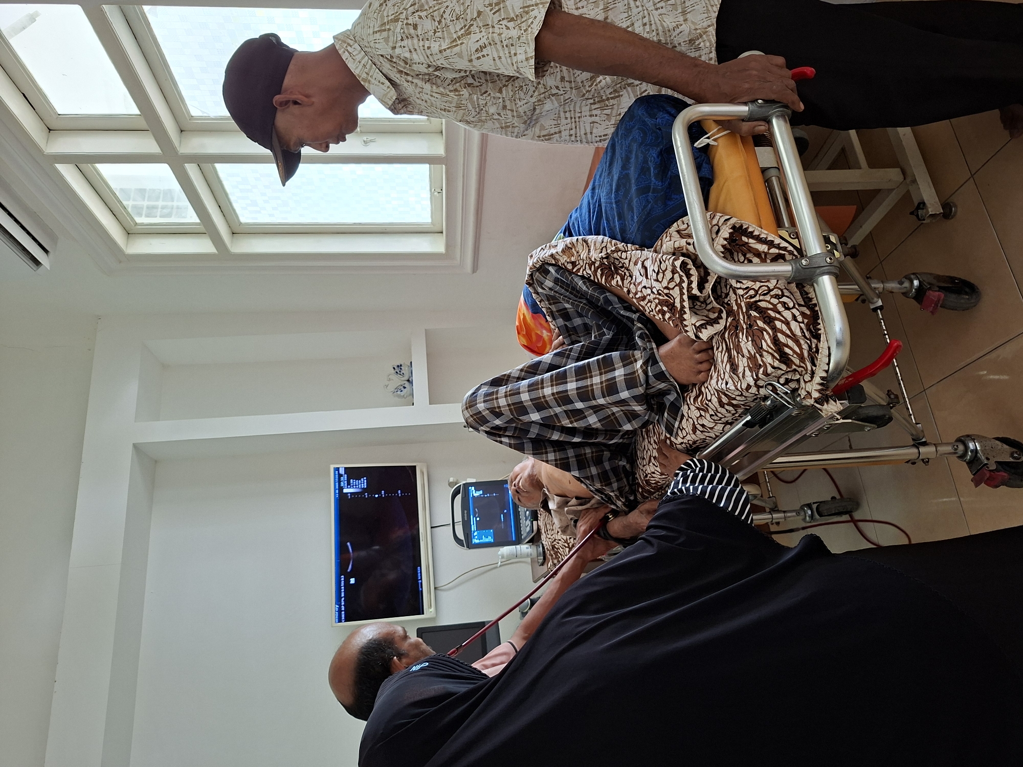 Location: Uleereuleung, Indonesia. An elderly person is undergoing an ultrasound examination in a medical room. A second person is assisting the patient and a third is performing the examination. Two ultrasound monitors are visible on the wall. A walker with wheels is near the patient. The room is tiled with white walls. A skylight is present.