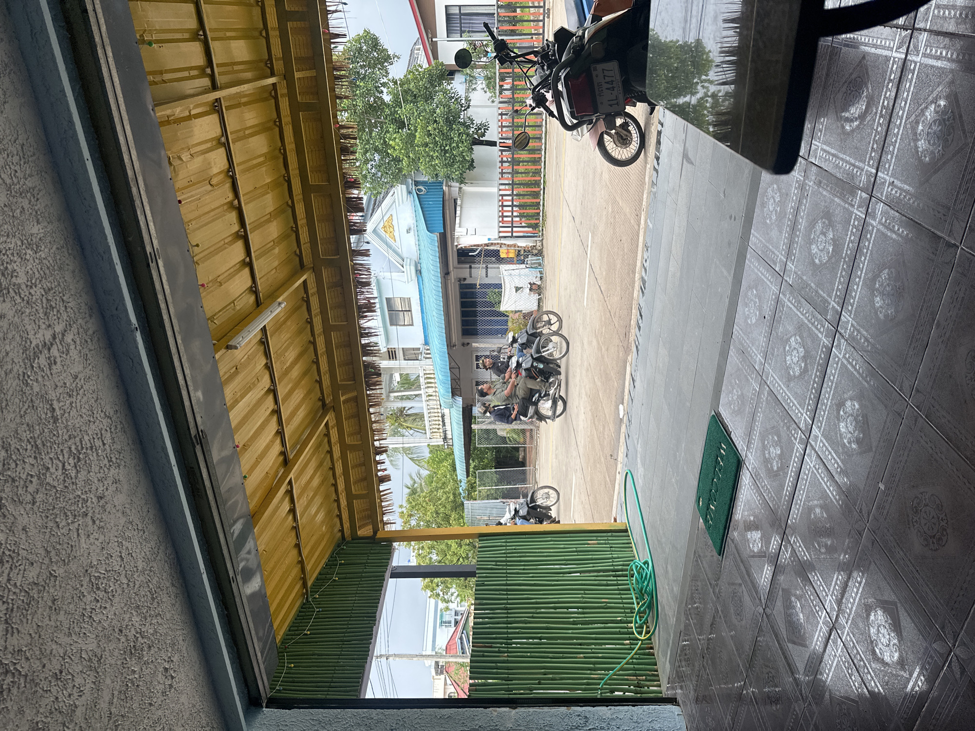 The image depicts a street scene in Kampot, Cambodia, viewed from what appears to be the interior of a covered outdoor seating area. The foreground is dominated by a tiled floor, with a green "Welcome" mat placed on it. To the left, a green garden hose is coiled on the pavement. Beyond this, a wall constructed of vertical bamboo poles forms a barrier.
The middle ground shows a paved street with several motorcycles. Three individuals are riding on two motorcycles moving away from the viewer. One motorcycle has two occupants, and another has one. On the right side of the street, a parked motorcycle is partially visible, with a license plate that reads "1L-4477 KAMPOT".
In the background, residential buildings are visible, characterized by light-colored facades and blue or teal roofs. A vibrant green tree stands between two of these buildings. A decorative fence, with alternating orange and black vertical bars, runs along the front of one of the properties. The sky is overcast, suggesting it may be a cloudy day. The overall impression is of a typical day in a residential area of Kampot.