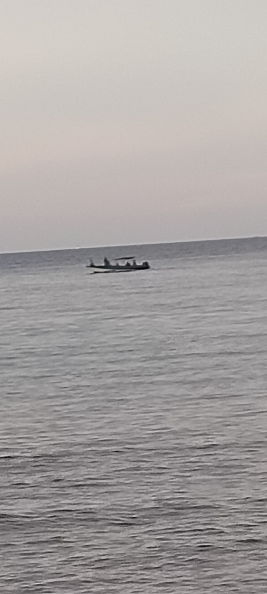 A long, narrow boat with a canopy over the back half is sailing on calm, gray water. The boat appears to be carrying several passengers, though they are not clearly discernible due to the distance and image quality. The horizon line is visible, separating the water from a pale, overcast sky. The scene is devoid of any specific landmarks or urban features, making it impossible to determine its precise location within Sabang, Indonesia, or any other city. The overall impression is one of a tranquil, possibly early morning or late afternoon, maritime setting.