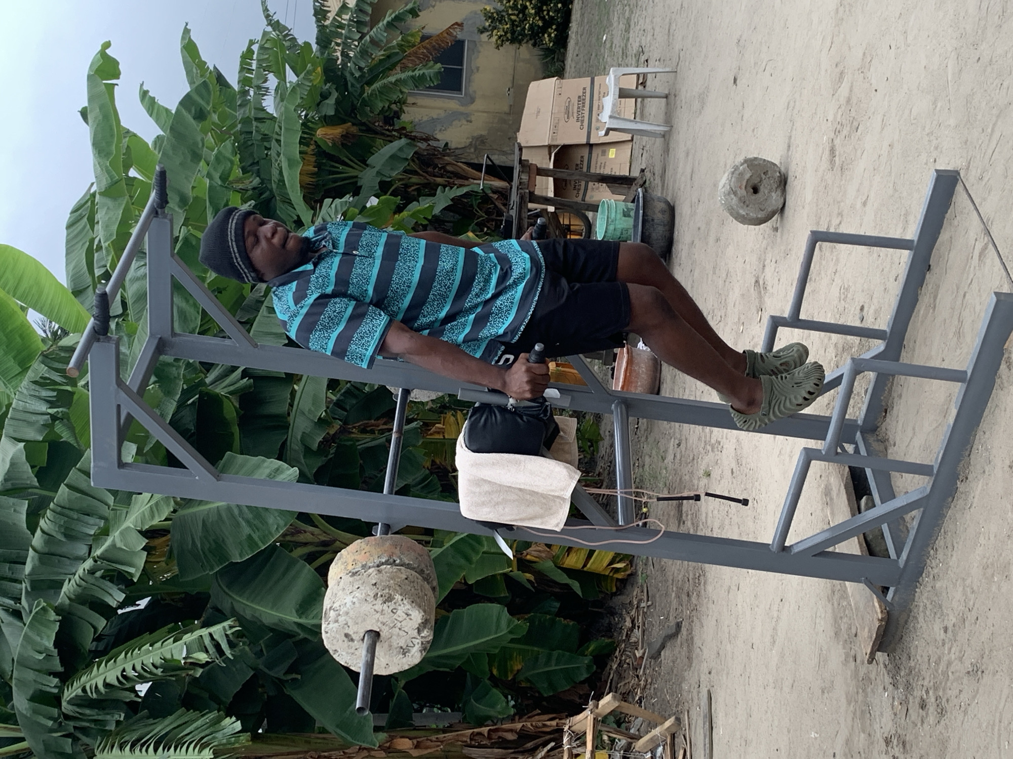 A Black man wearing a blue and black striped polo shirt, black shorts, and a black beanie is performing dips on a homemade dip station. The dip station is made of grey metal pipes and has a towel draped over one of the bars. On the left side of the dip station, there is a barbell with two large concrete weight plates. In the background, there are large banana trees, a building, and various objects on the ground including cardboard boxes, buckets, and a small stool. The ground is covered in dirt and sand. The scene appears to be an outdoor gym setup in a residential area of Ado, Nigeria.