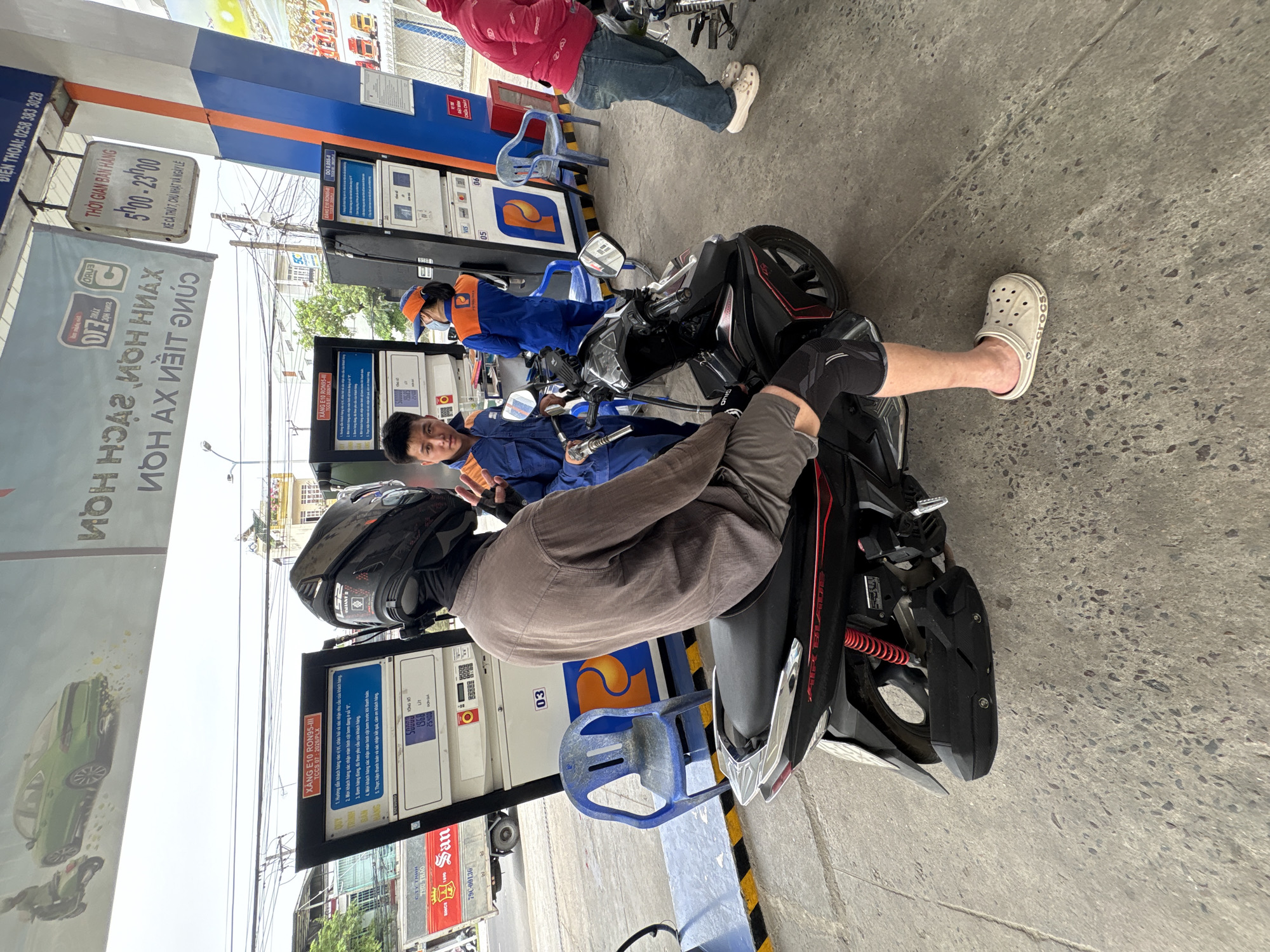 The image depicts a scene at a gas station in Bac Nha Trang Ward, Vietnam. Two men are interacting with a gas pump. One man sits on a motorcycle wearing a helmet and a grey shirt. His left leg is visible with black knee pads and his foot is bare except for a white Croc. The other man, dressed in a blue uniform with orange accents, stands near the motorcycle and appears to be filling the tank with gas. A second individual, also in blue uniform, stands next to a second pump further away. To the left, a sign displays the station's name, phone number, hours (5:00-23:00), and product information. There is another person wearing a red jacket and jeans sitting on a blue plastic chair near the farthest gas pump from the viewer. The ground is concrete. There are multiple blue plastic chairs. A truck can be seen in the background.
