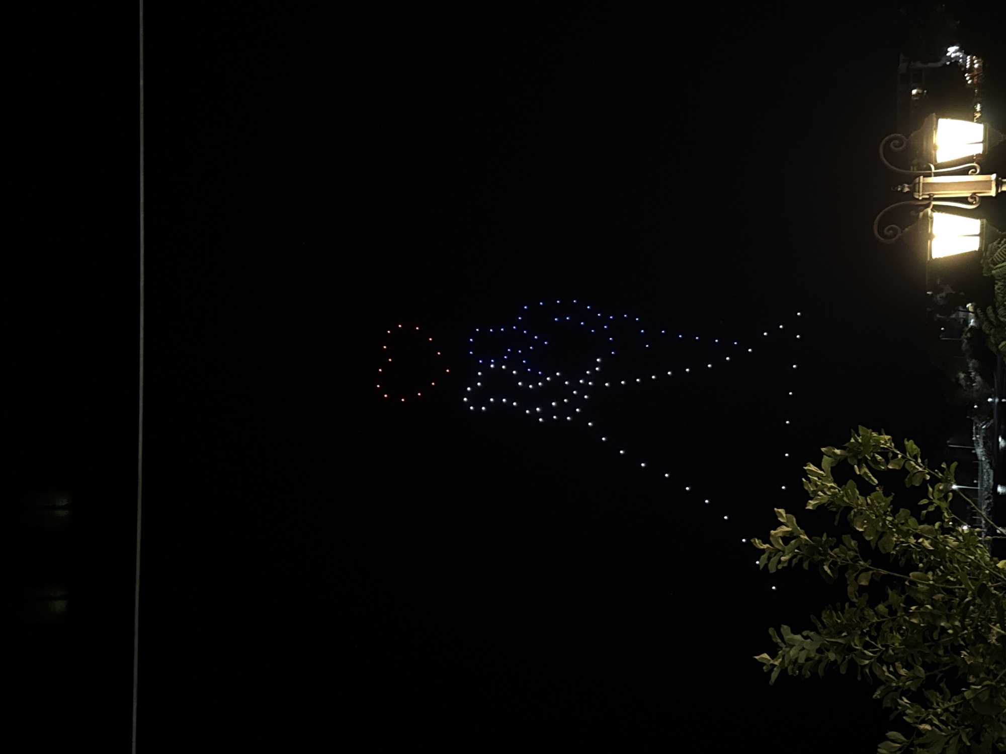 An aerial drone light show is taking place in the night sky above Tsarevo, Bulgaria. A silhouette of a person appears to be formed by numerous small lights, with a heart shape at the top of the figure. The drone formation is composed of white and blue lights, with a few red lights forming the heart. In the foreground, a tree with green leaves is visible on the left side of the frame, and a street lamp with two illuminated bulbs stands on the right. The background shows the dark sky with distant lights, suggesting a city or town below.