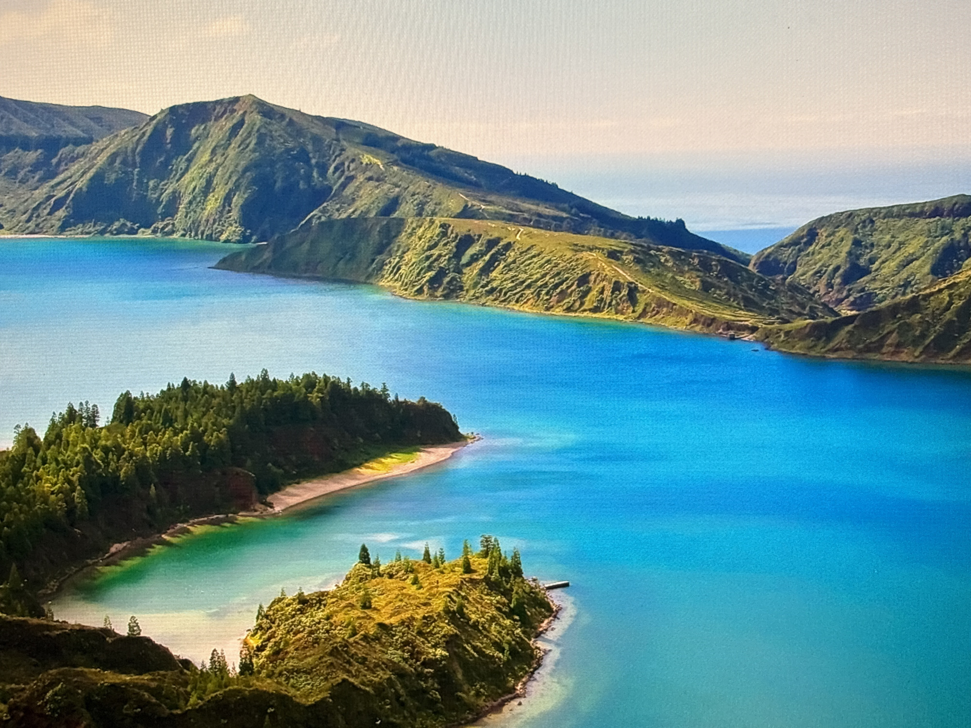 This is a landscape photograph of a vibrant blue lake surrounded by lush green hills and mountains. The water is a striking turquoise in some areas and a deeper blue in others, with clear visibility near the shore. The shoreline features a sandy beach and a small, tree-covered island with a pier. In the background, rolling hills rise steeply, covered in dense vegetation and showing signs of terraced paths. The sky is a pale, hazy blue. The image appears to be taken from a high vantage point, offering a panoramic view of the natural scenery. There are no people or man-made structures visible in the immediate vicinity, besides the small pier. This scene does not depict any location in the city of Sofia, Bulgaria.