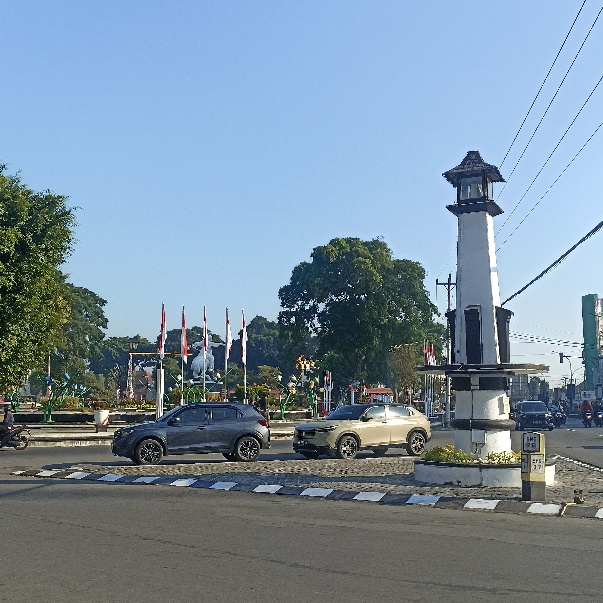 This is a daytime, eye-level shot of a roundabout in the city of Magelang, Indonesia. The foreground shows a paved road with a white and black striped curb. To the right, a tall, slender monument with a dark, pagoda-style roof stands prominently. Two cars are parked on the roundabout: a dark gray SUV and a light beige crossover. In the midground, flags with red and white stripes are displayed on poles, along with a large statue of a person on a horse. A dense green tree is on the left side of the frame, and other trees are visible in the background. Power lines crisscross the clear blue sky. Further back, traffic, including several motorcycles and cars, can be seen moving through the intersection. The overall scene depicts a typical urban intersection with public monuments and moderate traffic.