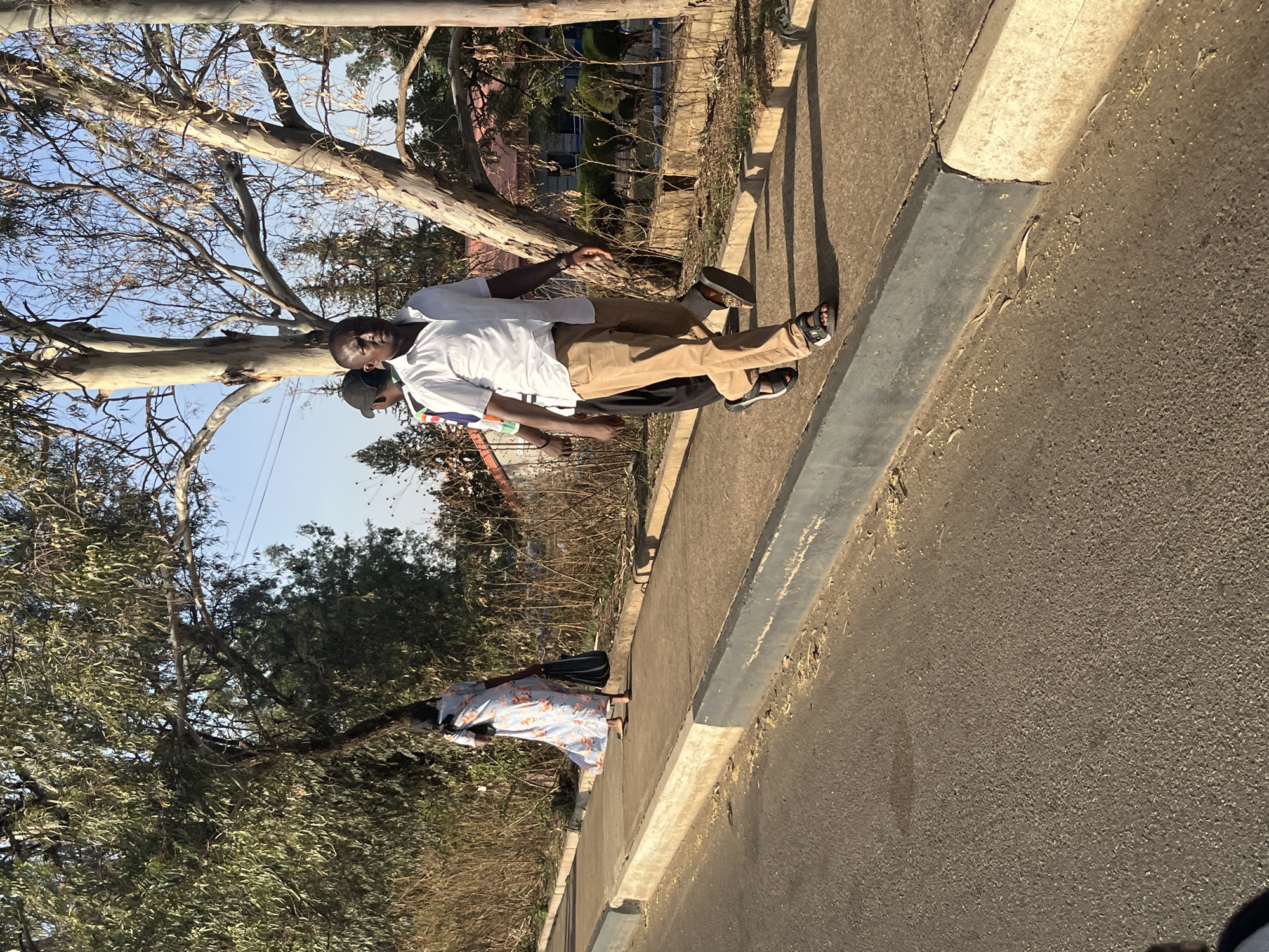 The image depicts three individuals on a road adjacent to a tree-lined curb in Gura Top, Nigeria. A man wearing a white shirt and khaki pants is walking with another man who is wearing a black jacket. A woman is walking behind them. The background includes a building and a fence. The road is paved. The sky is clear.