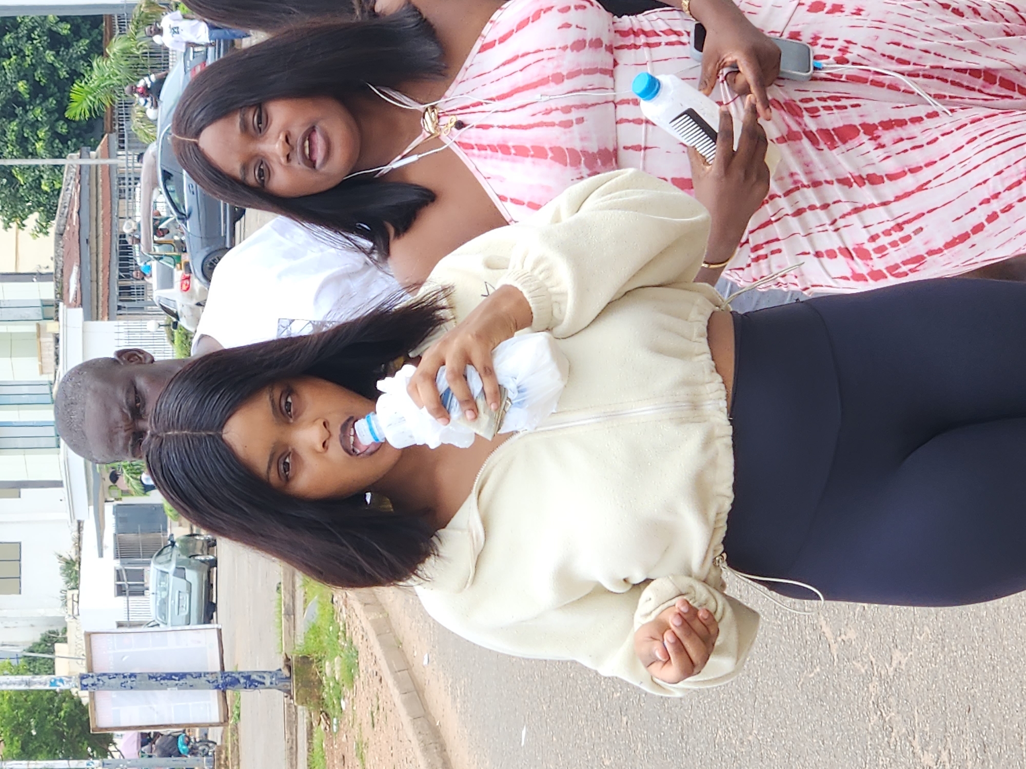This is a medium shot, eye-level, outdoor photograph taken in Jos, Nigeria. The image captures three individuals walking on a paved road. In the foreground, a woman with dark, shoulder-length hair is drinking from a plastic water bottle. She is wearing a cream-colored zip-up jacket over a dark-colored athletic top and leggings. To her right, another woman with similar dark hair is wearing a halter dress with a pink and white tie-dye pattern. She is holding a plastic water bottle in her right hand. Behind these two women, a man with dark skin and short hair is visible, looking towards the left of the frame. The background shows a street scene with cars, buildings, and trees, typical of a city environment. The lighting suggests it is daytime.