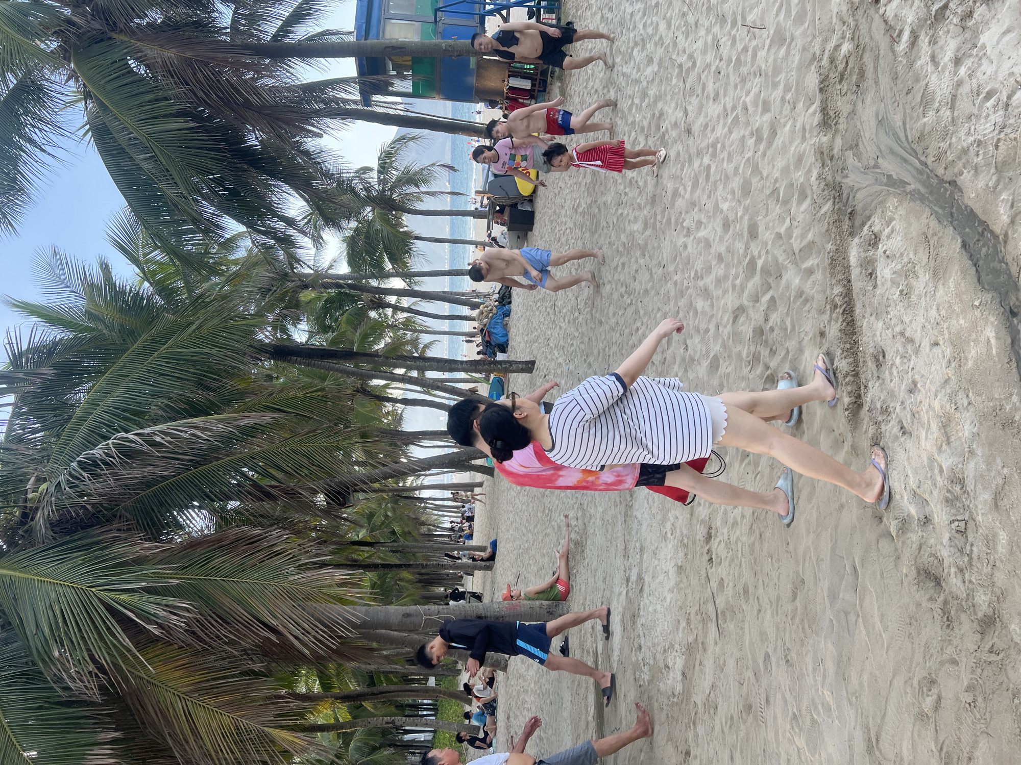 The image depicts a crowded beach scene in Đà Nẵng, Vietnam, under a bright, clear sky. Numerous palm trees line the sandy shore, casting shadows and providing a tropical ambiance. In the foreground, several individuals are visible, some walking and others relaxing.
On the left side of the image, a man in a white t-shirt and grey shorts is walking towards the viewer, his left leg bare. Beside him, a young man in a black hooded jacket and blue shorts walks away, looking at his phone. Further back, a person in a red shirt and black shorts is lying on the sand.
In the center of the image, a woman in a striped t-shirt and white shorts walks towards the right, her expression neutral. To her left, a man in a pink tie-dye shirt and black shorts stands. Behind them, a group of people are playing volleyball. A young girl in a red and white striped dress stands in the middle ground, facing forward. To her right, a man in red swim trunks and a young boy in blue swim trunks are walking. Further to the right, a man with a towel around his neck and black shorts stands, facing left.
In the background, more people are scattered across the beach, some sitting and others standing. A blue and yellow lifeguard tower is visible on the right side of the frame. The ocean is visible in the distance, with boats and mountains on the horizon. The sand shows footprints and some shallow channels, suggesting recent water flow.