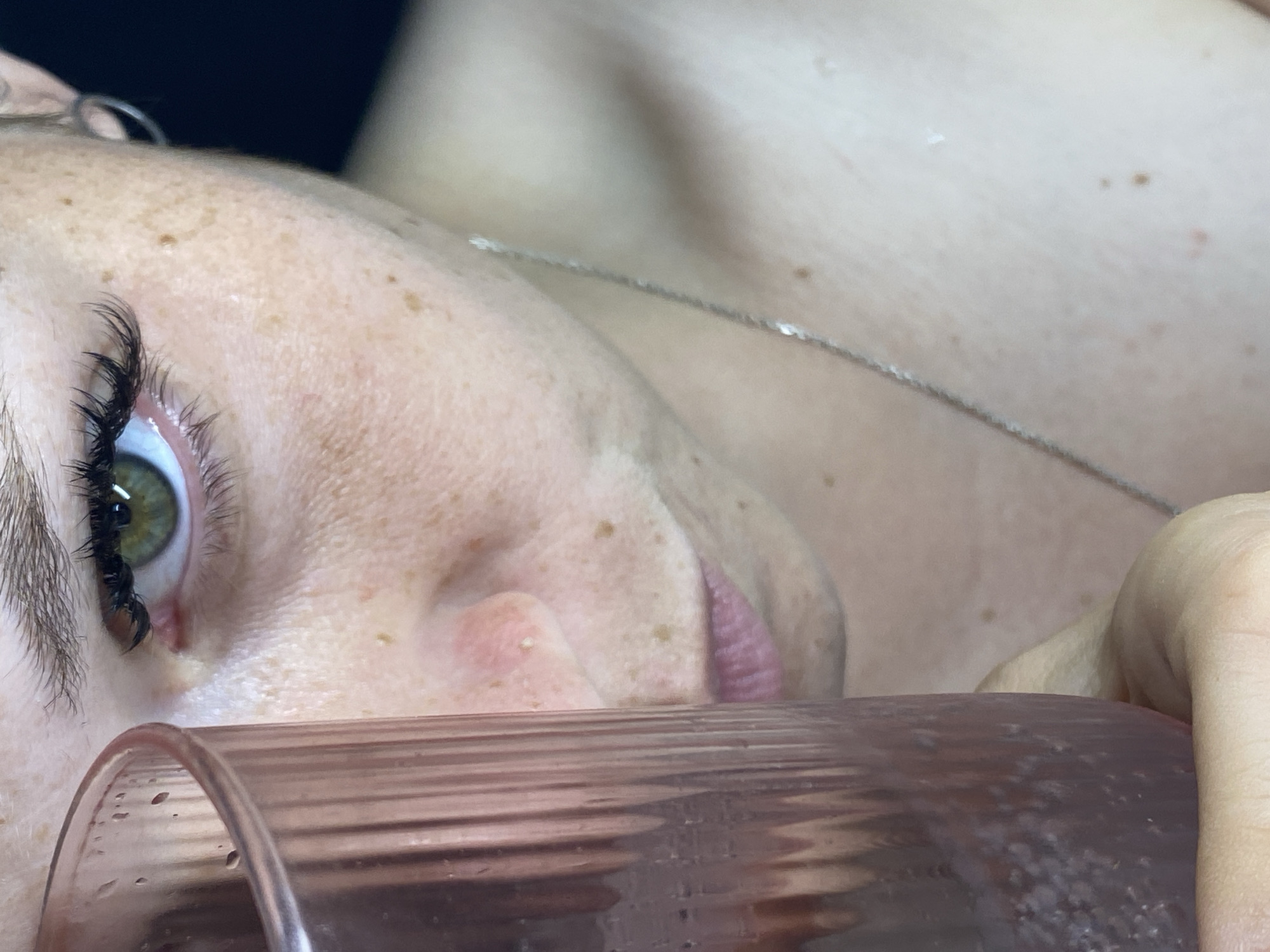 A close-up, chest-up shot shows a young person holding a pink, ridged glass in their left hand. The glass partially obscures their face from the left side. Their right eye, with green irises and long, dark eyelashes, is visible, looking directly at the camera. The person has fair skin with numerous freckles across their cheeks and nose. A thin silver necklace is visible on their collarbone. The background is dark and out of focus. The image does not contain any elements that would allow for determining a location in Chaikovsky, Russia.