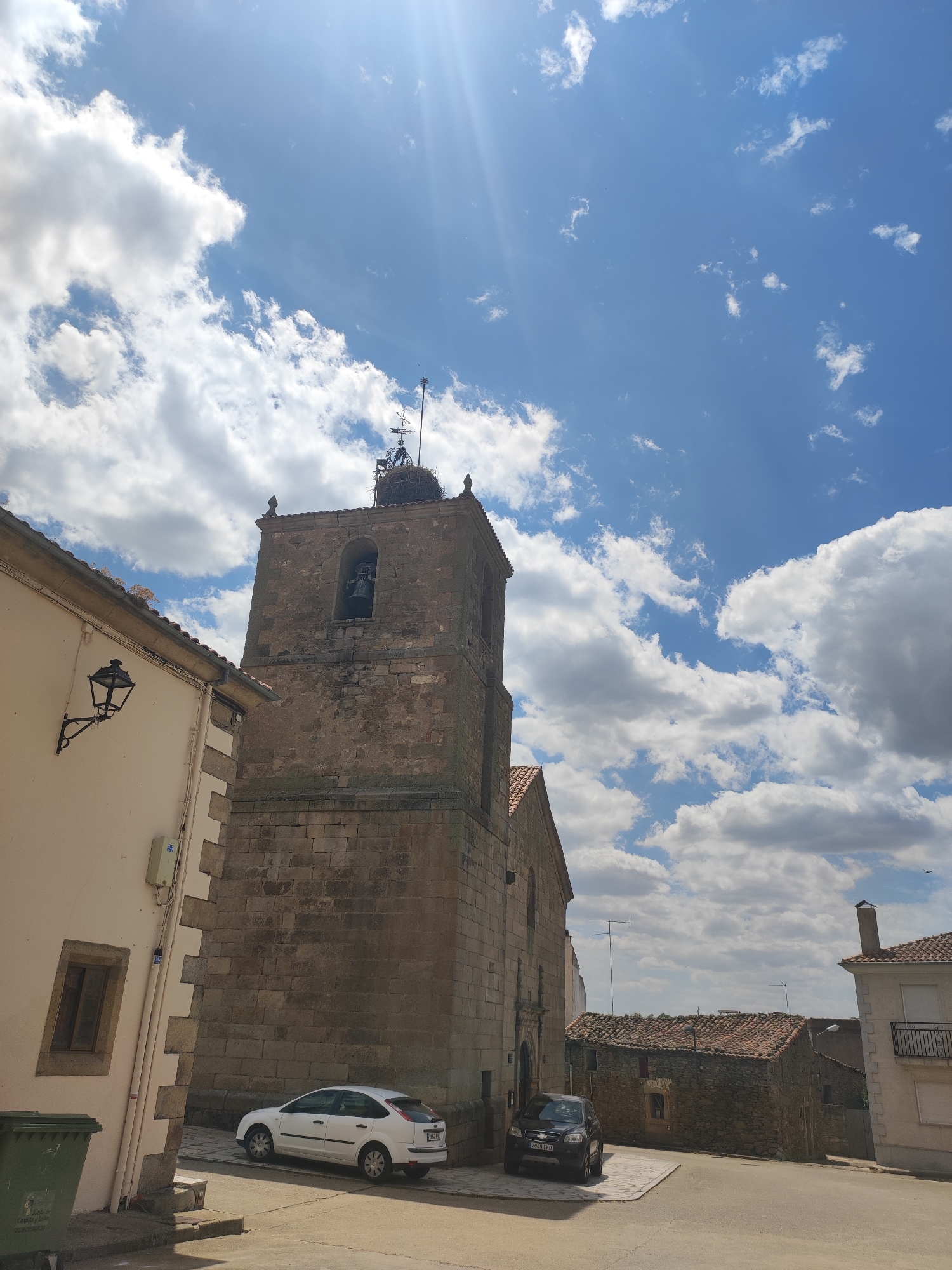 The image depicts a view of the town of Sobradillo, Spain, focusing on a church tower with a bell and a stork's nest at the top. The tower is constructed of rough-hewn stone blocks and has a weathered appearance. A large bell is visible within an arched opening in the tower. The sky above is a vibrant blue, populated by fluffy white clouds and illuminated by bright sunlight, with sun rays prominently visible. To the left of the tower, a section of a building with a cream-colored facade and a black lantern mounted on the wall is visible. The building features a corner constructed with contrasting lighter stone blocks and a small, recessed window with wooden shutters. A green garbage bin stands at the base of this building. In the foreground, on the street, a white car is parked. Further down the street, another dark-colored car is parked in front of a stone building with a red-tiled roof. The overall scene suggests a sunny day in a historic Spanish town.