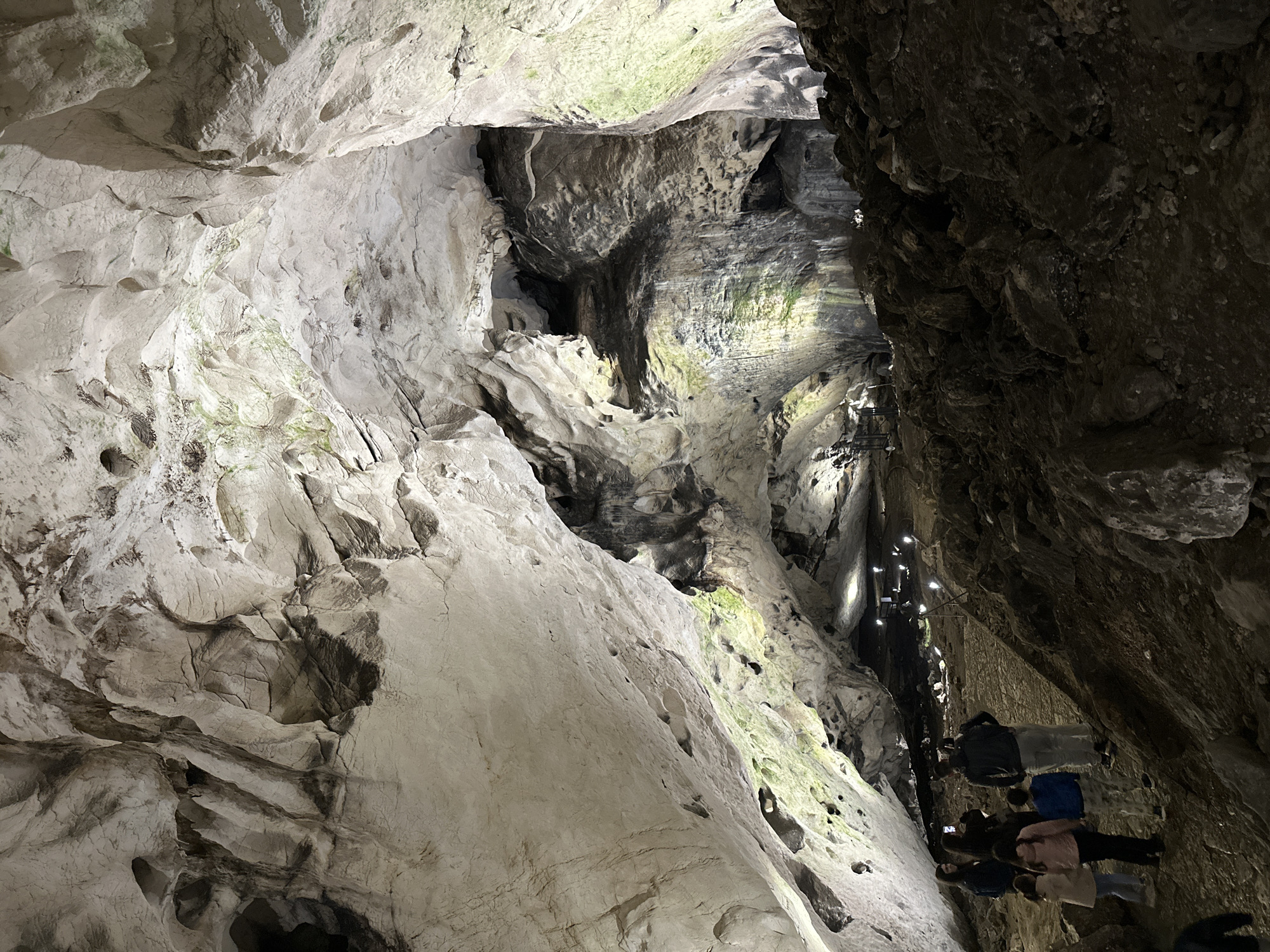 The image depicts a cave interior in Raianovtsi, Bulgaria. The cave features rock formations with varying textures and colors, ranging from light gray to dark brown. Some areas of the rock surfaces show signs of moisture or moss. A pathway with lights and a group of people are visible in the lower right portion of the image. The people are mostly dark, possibly due to the lighting conditions. The path seems to lead upwards through the cave.