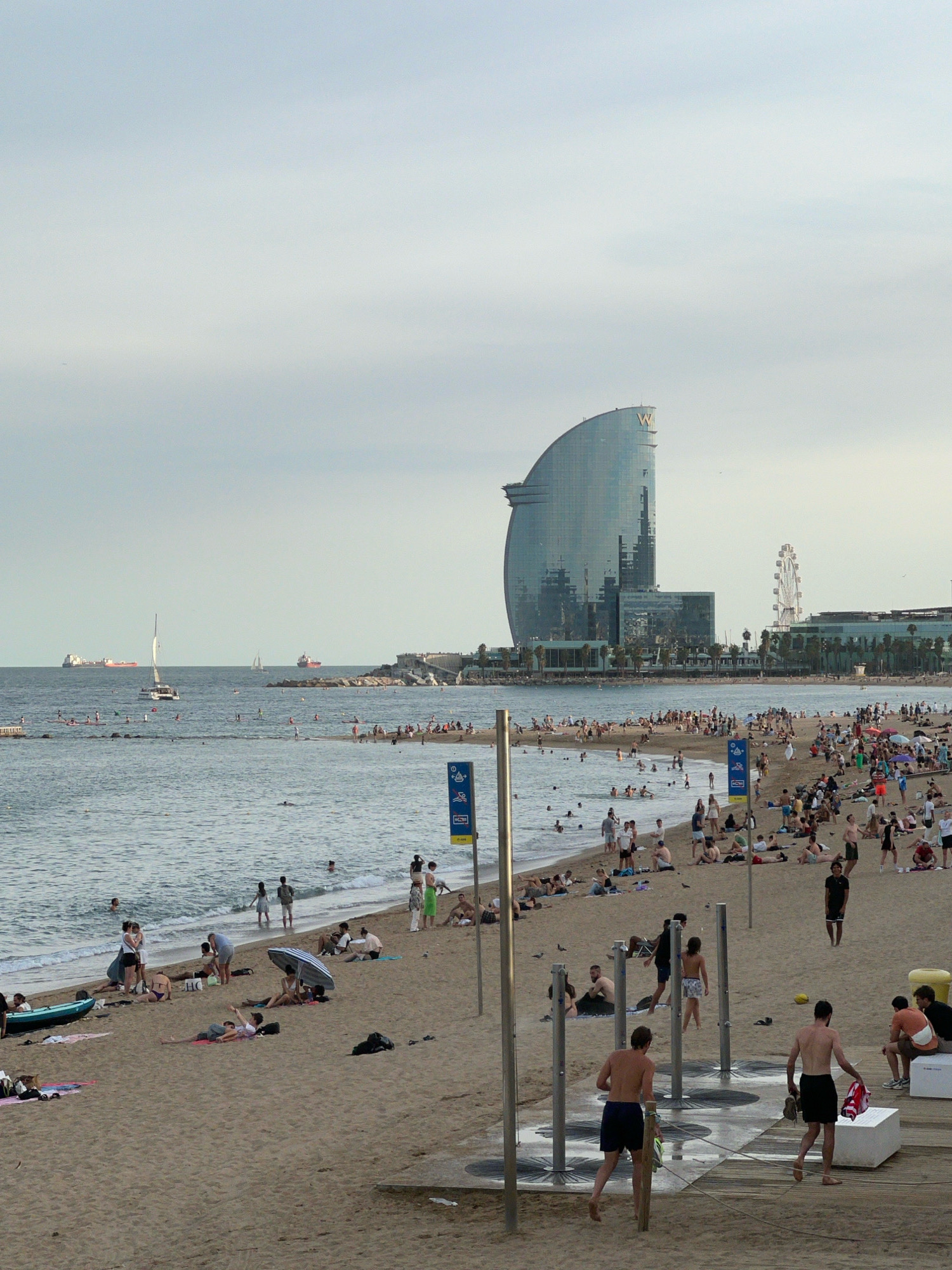 This image depicts a crowded beach scene in Barcelona, Spain, with the iconic W Hotel visible in the background. The beach is populated by numerous people sunbathing, swimming, and walking. Some are under umbrellas, while others are in the water or on the sand. In the foreground, there are beach showers, and several individuals are interacting near them. The sea is calm, with a few sailboats and cargo ships visible on the horizon. The sky is overcast, contributing to a soft, diffused light across the scene.