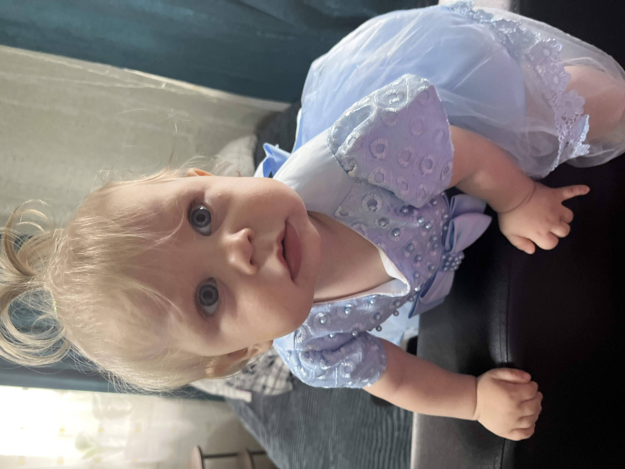 A baby with blonde hair styled in a high ponytail and blue eyes is crawling on a black surface, possibly a sofa or ottoman. The baby is wearing a light blue dress adorned with pearl-like beads and a bow. The background shows a room with a blue curtain and a blurred bed. The specific location in the city of Фоки, Russia, cannot be determined from the image alone.