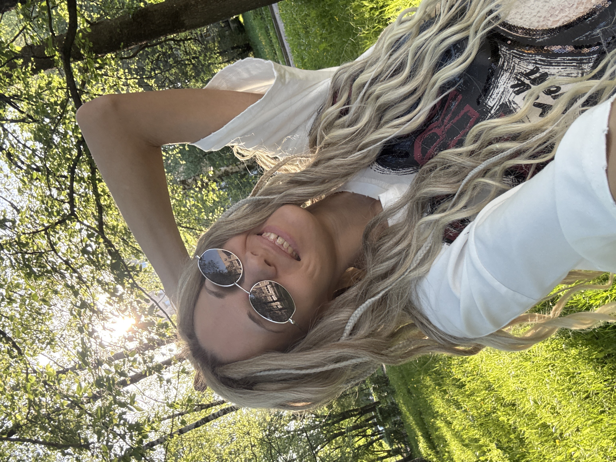 A woman with blonde hair, wearing round sunglasses, a white t-shirt, and a shirt with a graphic print, is lying on green grass in a park setting. The sun is shining through the trees, creating dappled light on the ground and her face. The sunglasses reflect buildings, likely in Rusanovo, Russia.