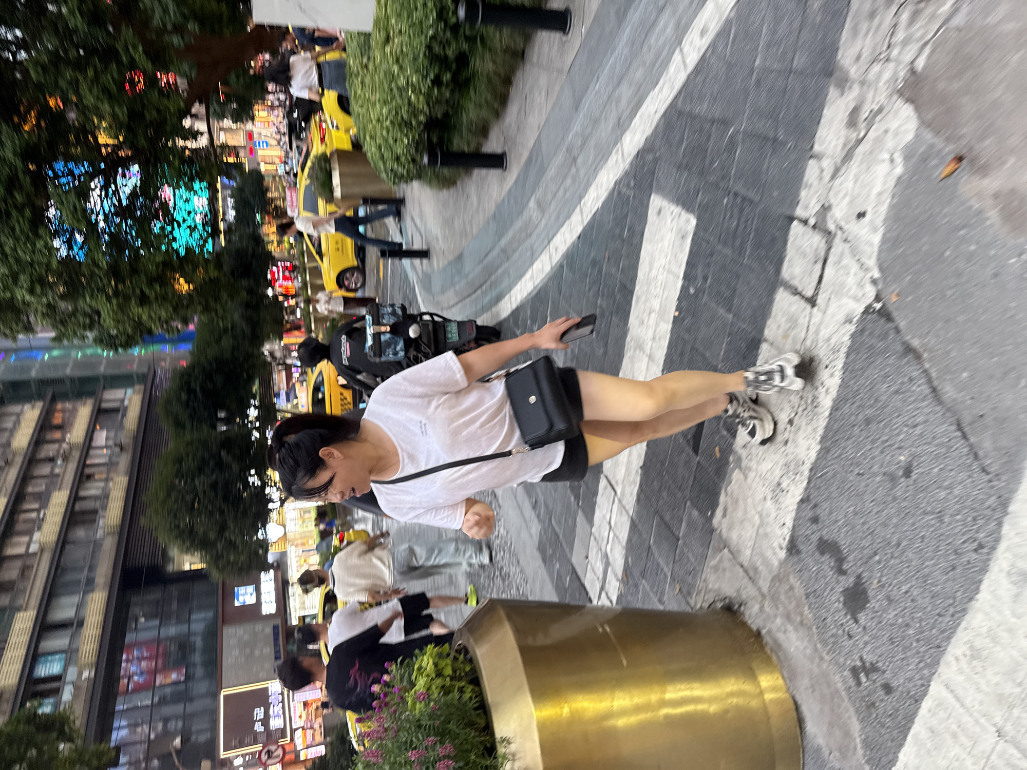 An outdoor, eye-level, medium shot captures a woman in motion in a city street in Yuzhong District, China. The woman, appearing to be of East Asian descent and in her 30s, is walking towards the right of the frame, her body angled away from the viewer. She has dark hair pulled back and is wearing a white t-shirt, black shorts, and athletic shoes. A black purse is slung across her body, and she holds a smartphone in her right hand.
The background depicts a bustling urban environment. Yellow taxis are visible, suggesting a commercial or transport hub. Buildings with multiple stories line the street, some adorned with illuminated signs. Green foliage from trees and planters is interspersed throughout the scene, adding a touch of nature to the concrete landscape. The ground is paved with distinct sections of gray and white, indicating a crosswalk or pedestrian pathway. The lighting suggests it is either late afternoon or early evening, with artificial lights beginning to illuminate the surroundings. The overall impression is that of a dynamic city scene with people engaged in everyday activities.