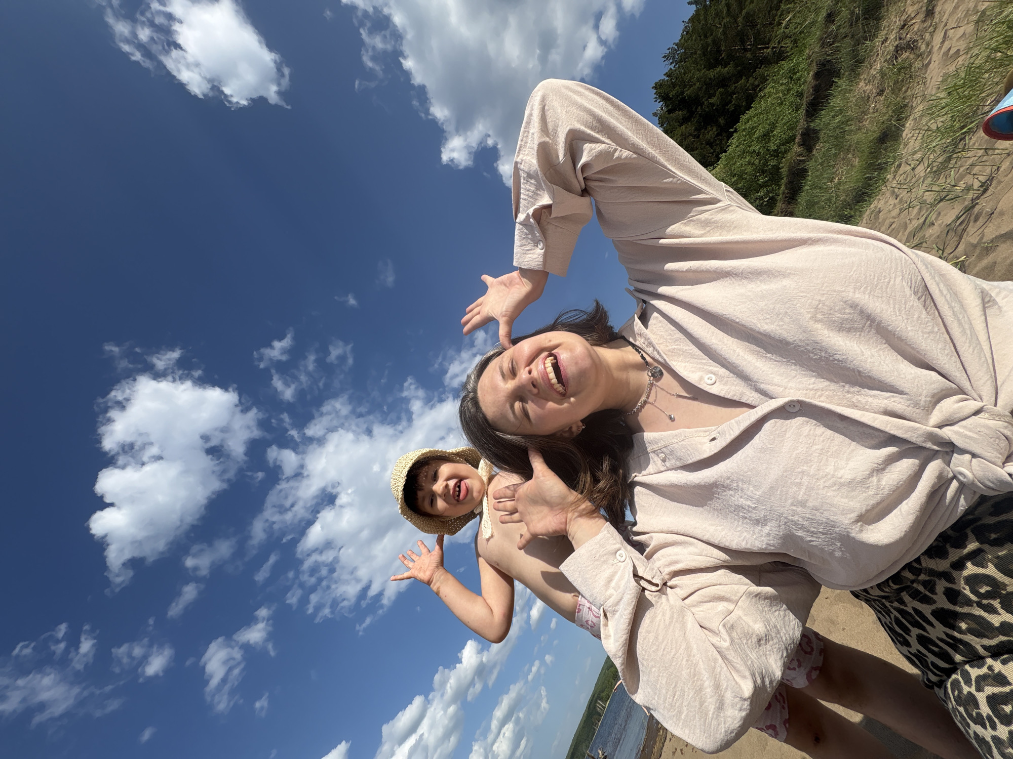 A low-angle shot shows two figures on a beach in the city of Chaikovsky, Russia, with a backdrop of a vivid blue sky punctuated by fluffy white clouds. The larger figure is an adult woman wearing a pale-colored button-down shirt and patterned shorts, posing with her arm raised and smiling broadly. A silver necklace is visible around her neck. The second figure, a young child wearing a straw hat, stands behind the woman. They are also posing with their arm raised and are partially obscured by the woman. A sandy beach stretches behind them and to the right, leading up to a grassy bank with trees visible in the top right corner. In the lower left corner, a body of water can be seen.
