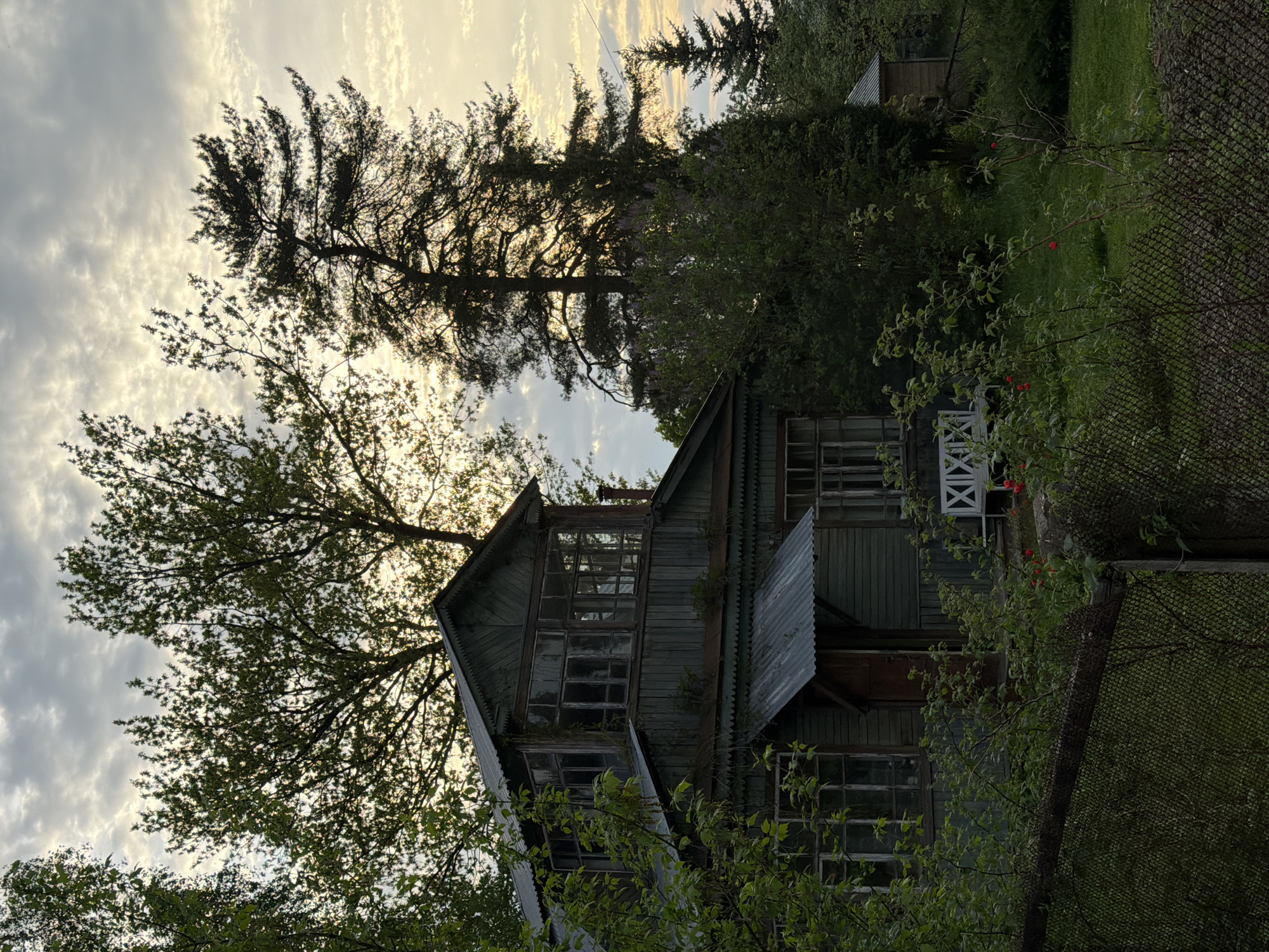 The image depicts a two-story wooden house with multiple windows. The house appears weathered, with visible signs of age and potential disrepair. The surrounding environment is lush, with trees obscuring parts of the house. A chain-link fence is visible on the right side of the image. The sky is overcast with clouds. The image is rotated. The location is Unknown, Unknown.