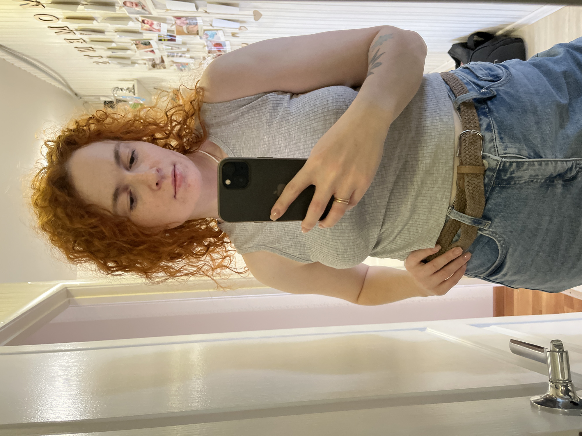 A young adult with fair skin and voluminous reddish-orange curly hair is captured in a mirror selfie, set in an indoor environment in Chaikovsky, Russia. The person, wearing a light grey ribbed tank top, blue denim jeans, and a woven brown belt with a silver buckle, holds a dark smartphone, likely an iPhone, in their right hand to take the photo. A simple ring adorns their right ring finger, and a subtle tattoo is visible on their upper left arm. Their expression is neutral and calm.
The reflection in the mirror reveals a brightly lit room. A white wall behind the person is decorated with numerous small photos and cards, suggesting a personalized display. Partially visible text on this wall, when read correctly through the reflection, appears to spell "KOFFEE" or a similar word, though it is slightly obscured. In the foreground of the reflected image, a clean white door with a modern silver handle is prominent. A dark backpack is faintly visible in the upper background. The overall scene conveys a casual, domestic atmosphere.