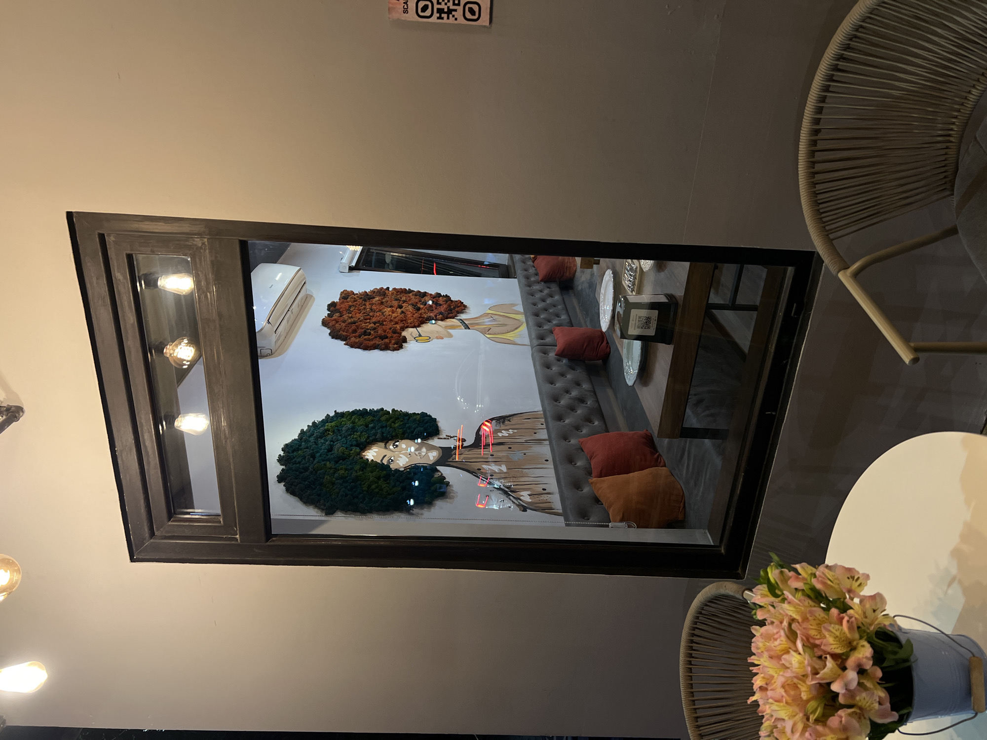 Captured indoors, the photograph showcases an art installation featuring two portraits of women with expansive afro hairstyles, one in earthy brown tones and the other in verdant green, viewed through a framed glass window. The installation sits inside a venue, potentially a cafe or restaurant, furnished with a grey tufted bench adorned with assorted pillows in terracotta and rust hues. Plates and objects are positioned on a nearby table, suggesting a dining or social setting. In the foreground, a vase containing a bouquet of light pink flowers rests on a white table, partially obscured by a woven chair. The scene gives the impression of a modern interior space, possibly a cafe or restaurant, that displays contemporary art. The presence of Armenian text, “Yerevan, Armenia” on the wall further narrows down the location.