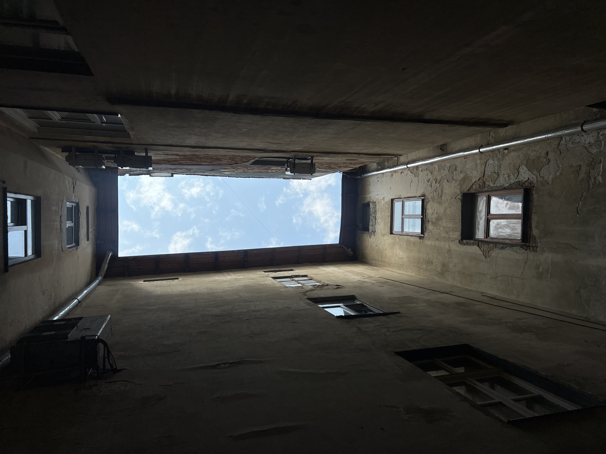 This is a low-angle shot looking up at the sky from a narrow courtyard in Lviv, Ukraine. The courtyard is flanked by tall, weathered buildings with multiple windows. The sky above is blue with scattered white clouds. A metal downspout runs vertically along one of the building walls. The overall impression is one of urban decay and enclosed spaces.