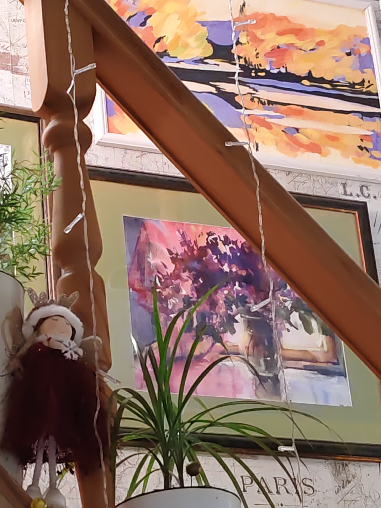 A festive holiday figurine of an angel with a white fluffy hat and red dress is placed on a wooden staircase railing. The angel holds a golden bell in its left hand. String lights are draped around the railing and the angel. In the background, several paintings hang on the wall, along with framed artwork and wallpaper. A green plant sits in a white pot to the right of the angel. The scene appears to be indoors, likely in a home in Rostov-on-Don, Russia, decorated for the holidays.
