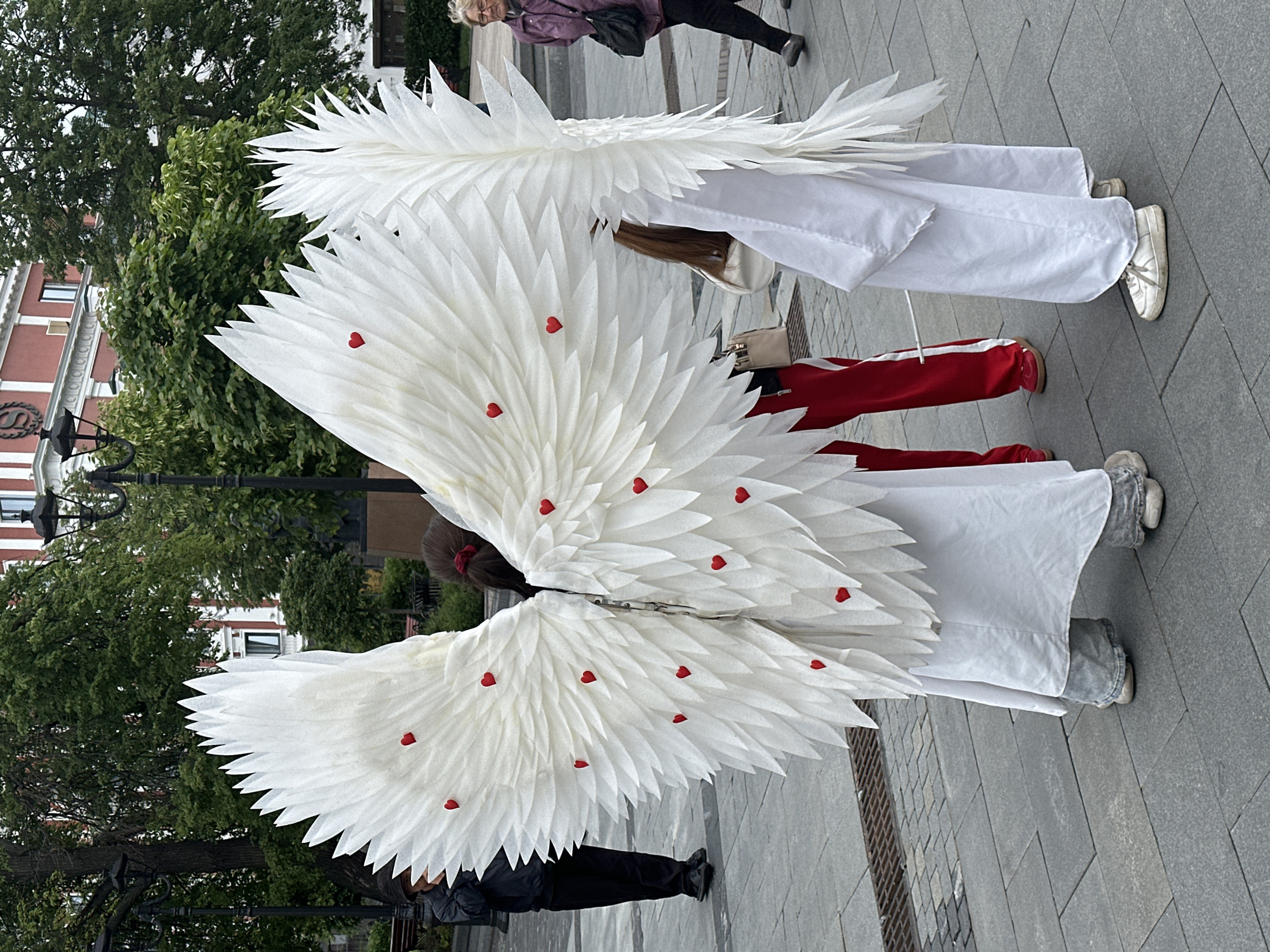 Two people dressed in large, white, feathery wings adorned with small red hearts are standing on a paved area in Nizhny Novgorod, Russia. They are wearing white robes. One person wears white sneakers, and the other has gray sneakers. The person in front is also wearing red athletic pants. Behind them, there are trees and buildings, including a building with a red facade. A street lamp is visible on the left side of the frame. Other people are walking nearby, with one visible person wearing a purple jacket.