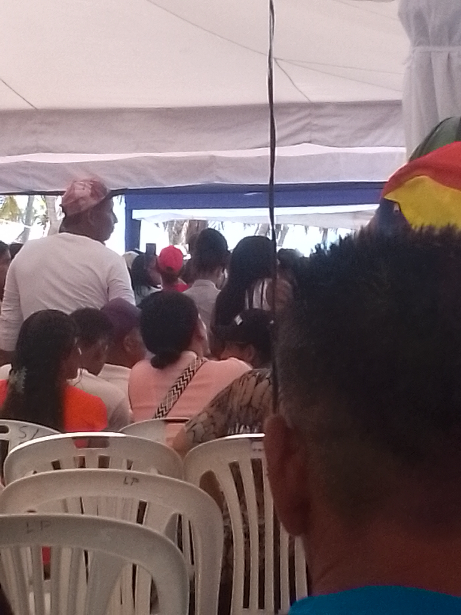 The image shows a crowd of people seated in white plastic chairs under a white canopy in what appears to be an outdoor event. The location is identified as Juan Griego, Venezuela. The crowd is diverse, with people of different ages, genders, and ethnicities. Some are wearing casual clothing, while others are dressed more formally. The chairs are arranged in rows, and the people are generally facing forward. The lighting is bright, suggesting it is daytime. The focus of the image is slightly blurred, but the general scene is discernible. There are some people standing in the background.