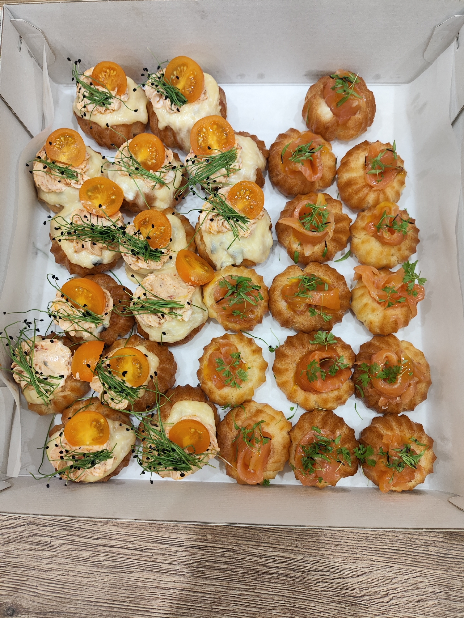 A top-down, medium shot shows a white cardboard box filled with an assortment of small, savory pastries, likely hors d'oeuvres or appetizers. The pastries are arranged in neat rows. Some are topped with a dollop of pinkish-orange filling, a slice of bright orange cherry tomato, and delicate green sprigs, possibly chives or microgreens, along with black seeds. Other pastries are adorned with thin strips of smoked salmon and a few sprigs of green herbs. The box is placed on a light brown wooden surface with a distinct grain pattern, reminiscent of a table. The overall lighting is bright and even, suggesting an indoor setting. There are no people or discernible actions in the image. The scene is static and focused solely on the presentation of the food within the box.
