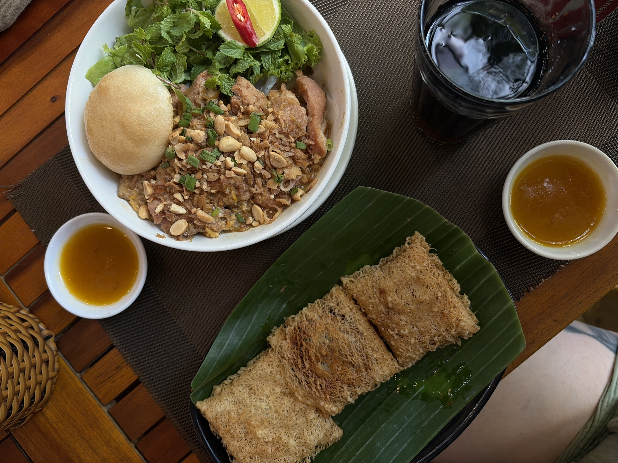 An overhead shot captures a Vietnamese meal served on a wooden slatted table. The scene features a main dish in a large white bowl, a side dish on a black plate with a banana leaf, two small bowls of dipping sauce, and a glass of dark beverage.
The main dish consists of stir-fried or grilled meat, likely pork or chicken, coated in a savory sauce and generously topped with chopped roasted peanuts and green onions. It is accompanied by fresh green herbs, including mint and lettuce, and garnished with a lime wedge and a red chili slice. A small, round, plain fried bun is also present in the bowl.
The side dish, placed on a vibrant green banana leaf, consists of three rectangular, crispy, net-like items, characteristic of *chả giò rế* (net spring rolls) or a similar fried dish common in Vietnam. A small amount of green liquid, possibly a sauce, is visible on the banana leaf.
Two small white bowls contain a yellowish-orange dipping sauce, likely a fish sauce-based condiment. A tall glass holds a dark, iced beverage, such as cola or iced tea.
The meal is set on a rustic wooden table, with a dark brown woven placemat beneath the main bowl and the drink. A woven basket is partially visible on the left. Part of a person's leg, wearing light-colored shorts or a skirt and featuring a tattoo on the thigh, is visible in the bottom right corner. The bright lighting suggests the meal is taking place during the day in Đà Nẵng, Vietnam. No discernible text is visible in the image.