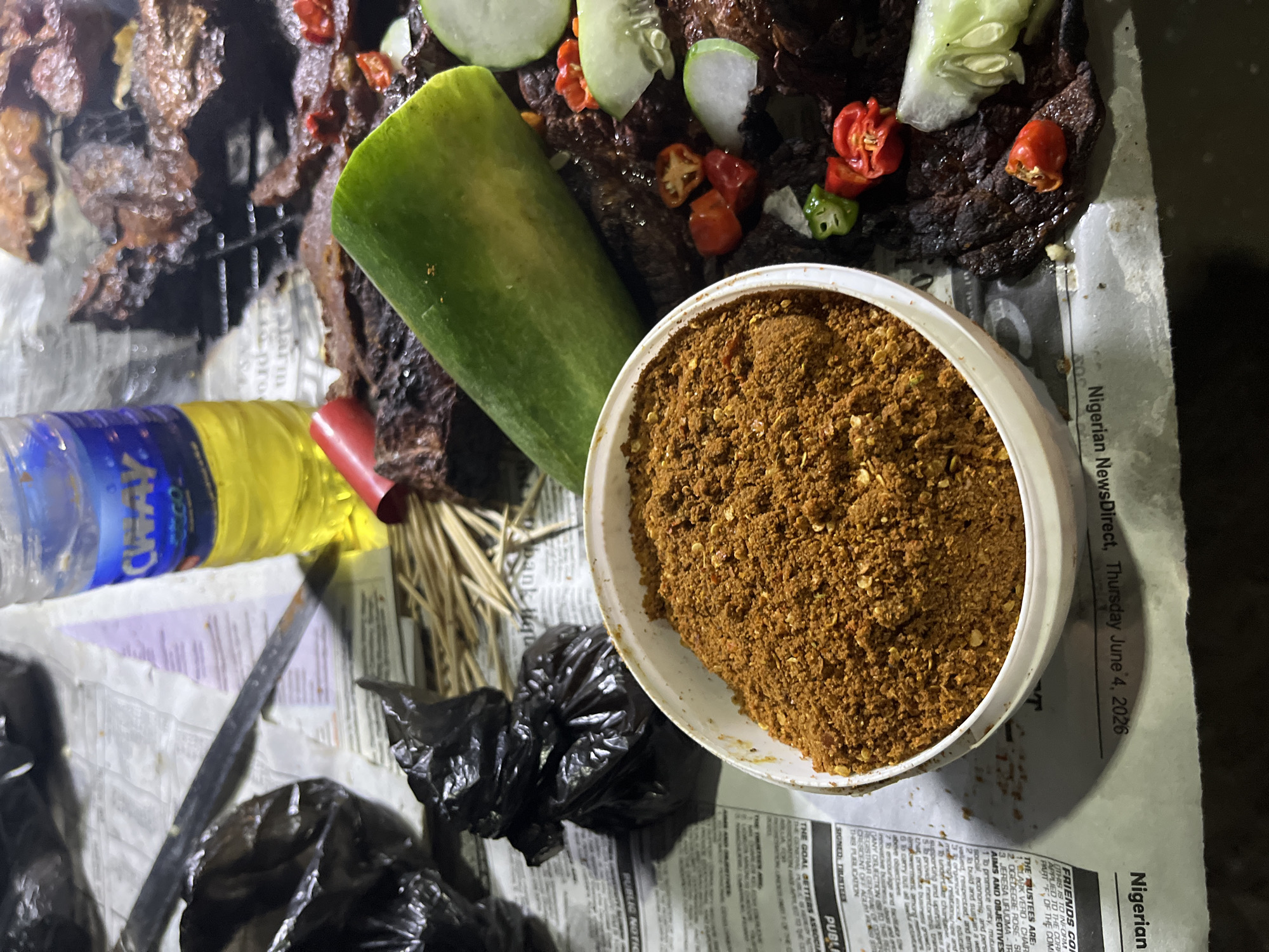 A close-up, overhead shot captures a collection of food items laid out on a surface covered with newspapers. In the foreground, a white plastic bowl is filled with a coarse, reddish-brown spice blend, possibly pepper or a seasoning mix. To the right of the bowl, several pieces of dark, grilled meat are visible, some with slices of cucumber and small, red and green chili peppers scattered amongst them. A large, partially peeled cucumber sits prominently above the meat. Behind the food, a partially filled bottle of "CHIVAS" branded liquid, likely water, is present, along with a red object, possibly a piece of food or a utensil. Two black plastic bags are also visible, one appearing to be a trash bag. The newspapers beneath the food are from "Nigerian NewsDirect," dated Thursday, June 4, 2026, with visible headlines and text indicating a local publication. The setting appears to be an outdoor or informal food stall, potentially in Abuja, Nigeria, given the newspaper source. The lighting is dim, suggesting an evening or nighttime scene.