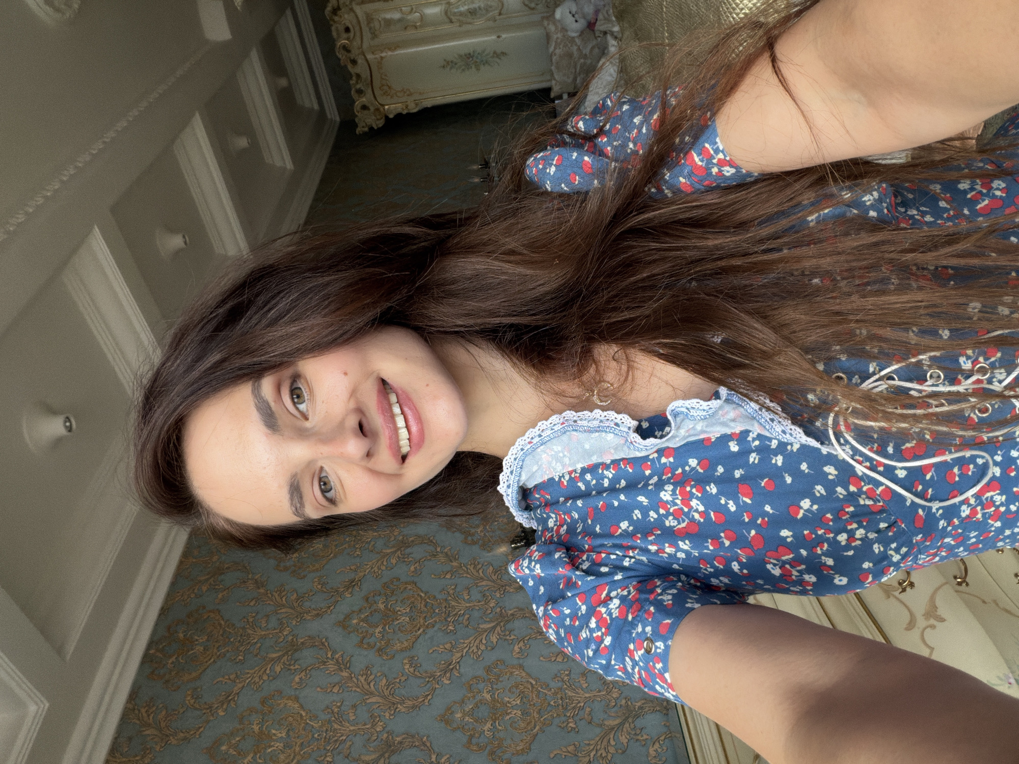 In this selfie, a young woman with long, brown hair smiles at the camera. She is wearing a blue dress with a red and white floral print and a white lace trim around the neckline. Her eyes are light brown, and she has a smattering of freckles across her nose and cheeks. Her hair is long and wavy, falling around her shoulders and down her back. The background of the image is a richly decorated room, featuring patterned wallpaper and ornate furniture, suggesting a luxurious indoor setting. The lighting appears to be natural, possibly from a window, casting a soft glow on her face and hinting at daytime. The overall mood is relaxed and cheerful. The location context provided is Ufa, Russia, though there are no explicit visual cues in the image to confirm this. No text is visible in the image.