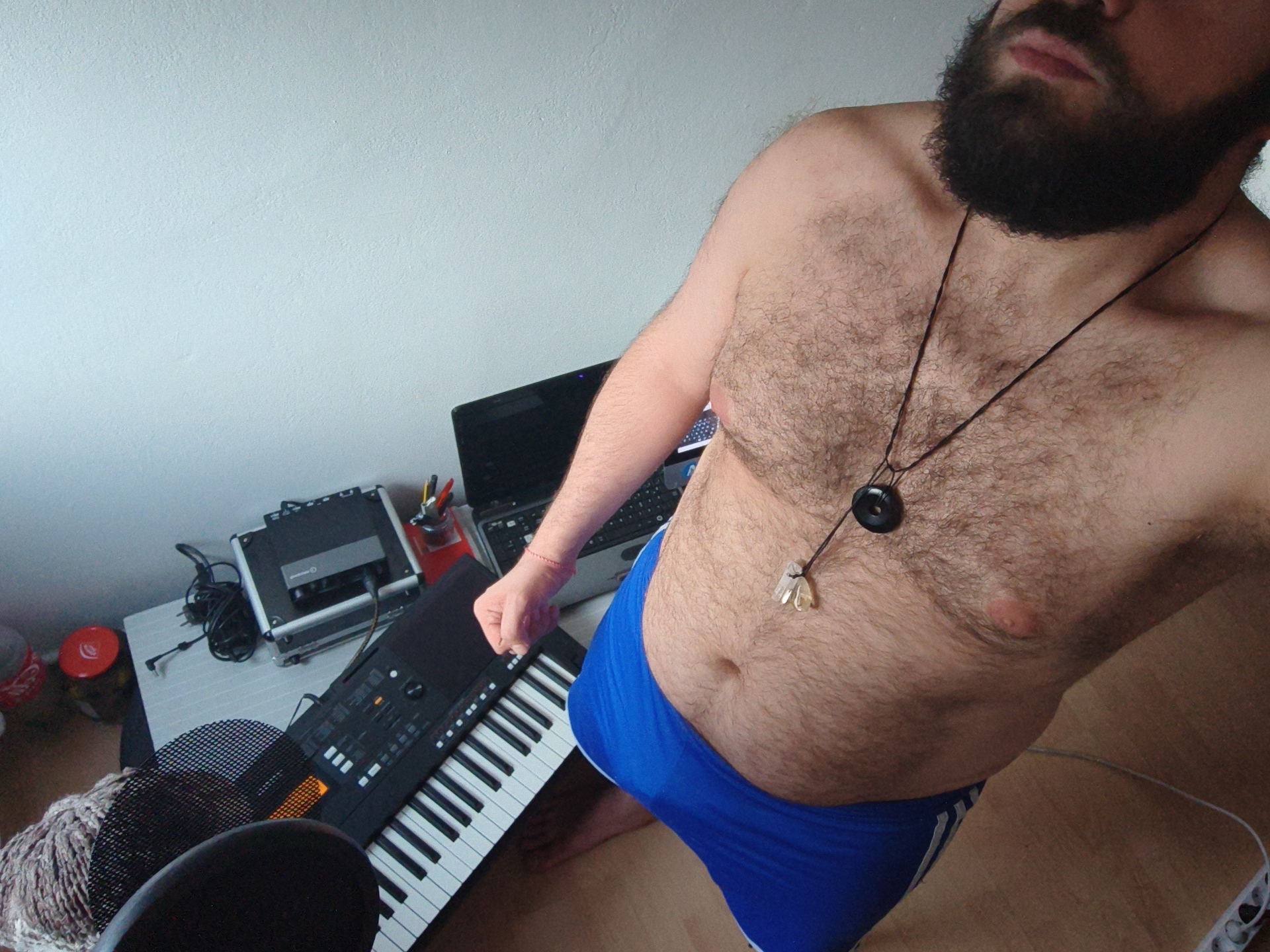 A high-angle shot depicts a fair-skinned, hirsute male with a dark beard, wearing blue athletic briefs, positioned in a room featuring a keyboard, laptop, microphone, and other equipment. The environment suggests a recording or music production space. The location, Diepholz, Germany, is assumed based on the prompt's instructions, but is not visually verifiable within the image.