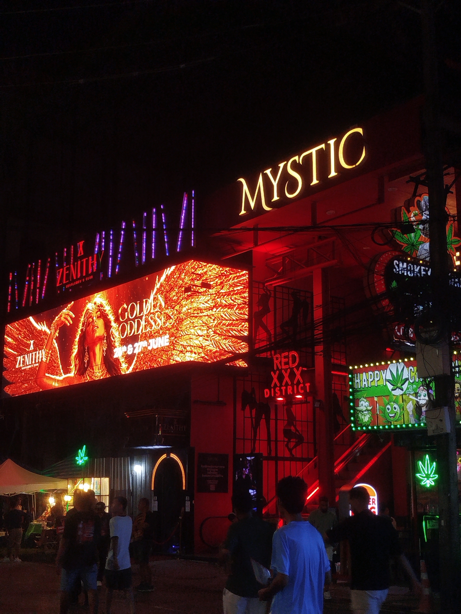 An outdoor nighttime scene displays several neon signs and illuminated advertisements. A large, horizontal LED screen on the left features an image of a woman with wings and the text "ZENITHY GOLDEN GODDESSES 24th & 27th JUNE." Above this screen, a smaller sign reads "ZENITHY" with purple and blue lights. To the right, a large, illuminated sign displays the word "MYSTIC" in yellow. Further to the right, there are more signs with green and red neon lights, including depictions of cannabis leaves and cartoonish characters. One sign reads "RED XXX DISTRICT." A few people are visible in the foreground and background, some walking and others standing. The environment appears to be a street or entertainment district, with the overall lighting dominated by the colorful neon signs.