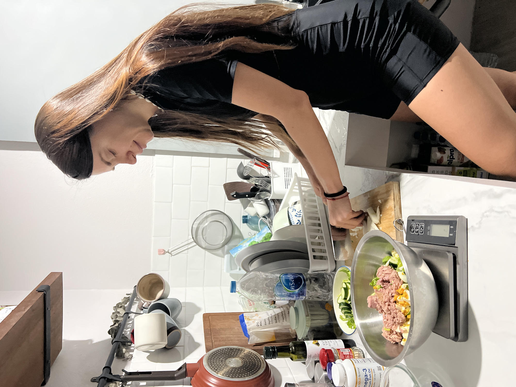 A woman in a black top and shorts is cutting food on a wooden cutting board in a kitchen. Next to her is a stainless steel bowl filled with tuna, lettuce, and hard-boiled eggs, placed on a digital kitchen scale. Various kitchen items and food containers are scattered on the countertop, including mugs, a strainer, a frying pan, bottles of oil, and several containers of supplements and ingredients. The environment includes white tiled walls and cabinets. It's not possible to identify the specific location as being in the city of บ้านบางรักษ์, Thailand, from this image alone.