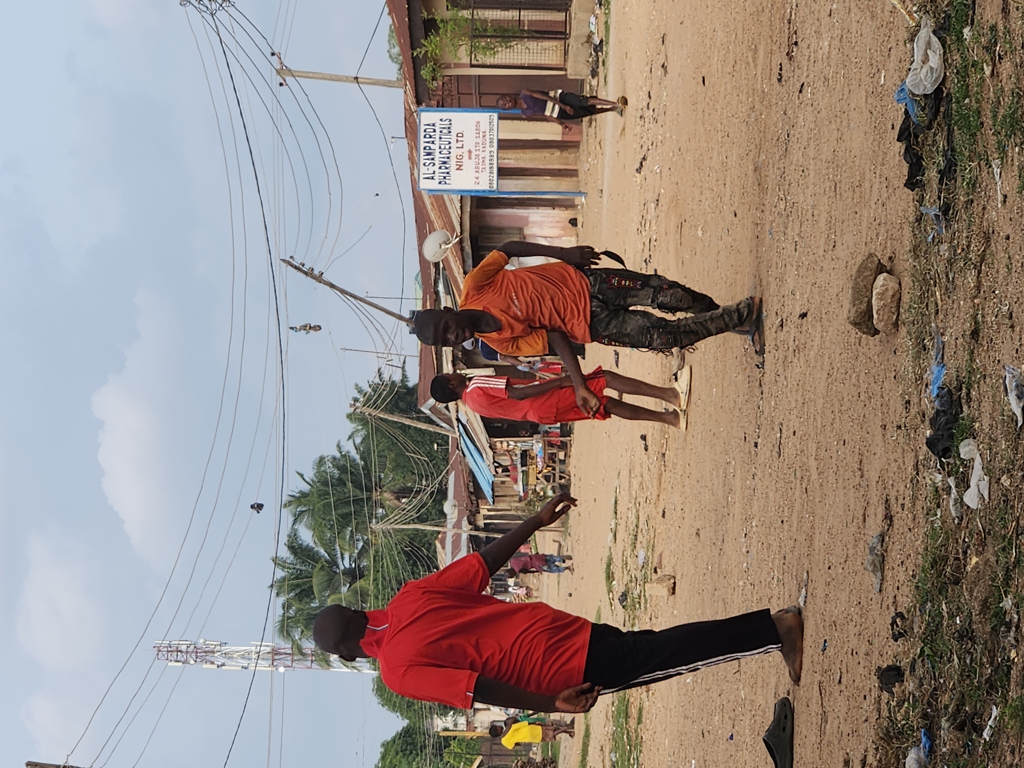 A street scene unfolds in Sabon Tasha, Nigeria, characterized by a dusty, unpaved road and modest, tin-roofed buildings lining one side. Overhead, a complex network of electrical wires crisscrosses the sky, supported by utility poles and a prominent cell tower in the distance.
In the foreground, a young man in a bright orange t-shirt and patterned pants is captured mid-stride, his body angled as if engaged in an activity or conversation. To his left, a man in a red polo shirt and black pants faces away from the camera, his left arm extended, also appearing to be interacting with someone or something outside the frame. Further down the street, other individuals are visible, including a person in a yellow shirt and others in red clothing. One person, a boy in a purple and white striped shirt, walks toward the right side of the image. The ground is littered with debris, including plastic bags and other refuse, suggesting a somewhat neglected environment. A blue sign with white lettering, identifying "AL-SAMPARDA PHARMACEUTICALS NIG. LTD." and an address on "ABUJA STR. SABON TASHA KADUNA," stands prominently near the buildings. The sky is cloudy, with patches of blue visible.