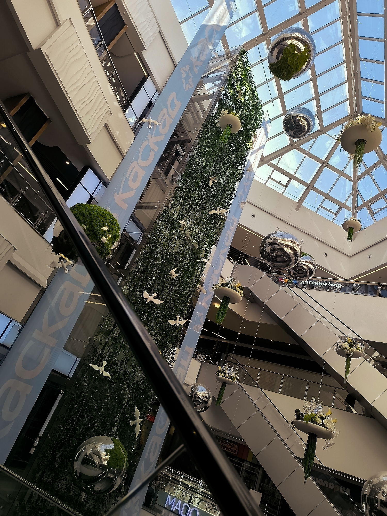 The image depicts the interior of a multi-level shopping mall, characterized by escalators, glass railings, and a large glass ceiling allowing natural light to filter in. Hanging from the ceiling are several large, reflective spherical ornaments and decorative floral arrangements suspended from white, bowl-like structures. A tall, vertical green structure, possibly a decorative tree or wall, is centrally located, adorned with numerous white bird figurines. Large banners with blue lettering are visible on the sides of the central structure, one of which appears to have the Cyrillic text "каскад" (kaskad). The overall impression is one of a modern, well-lit commercial space with a focus on decorative elements and a sense of openness. The location is identified as being in the city administration of Qarağandı, Kazakhstan, suggesting this is a prominent shopping center within that region. There are no discernible individuals in the image, focusing the composition on the architecture and decorations.