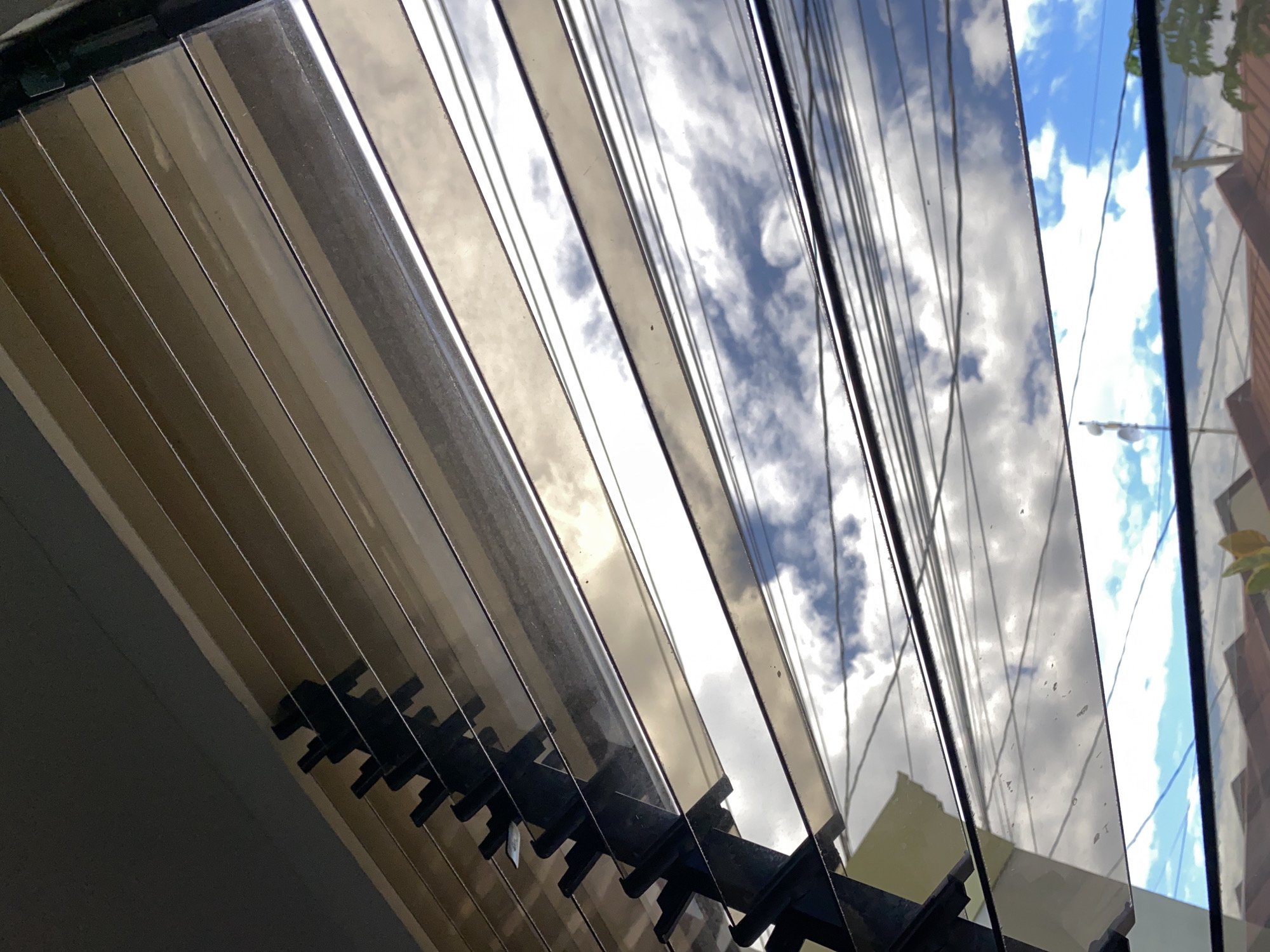 The image is a low-angle, close-up shot of louvre windows seen from inside a building, looking outwards. The louvers themselves are wide, translucent, and reflect the sky and surroundings. Through the gaps between the louvers, a bright sky with scattered clouds is visible. The clouds vary in texture and density, with some appearing white and fluffy and others darker and more ominous. Several power lines are strung across the view, creating a complex network of dark horizontal lines against the sky. In the distance, the roofs of houses are visible, some with terracotta tiles. A portion of a light-colored building is also seen on the left side of the frame. The overall impression is one of an urban or suburban environment, likely with a tropical climate given the bright sky and abundance of clouds. The specific location of Davao City, Philippines, suggests a warm and humid atmosphere, which would be consistent with the appearance of the sky and the types of houses visible.