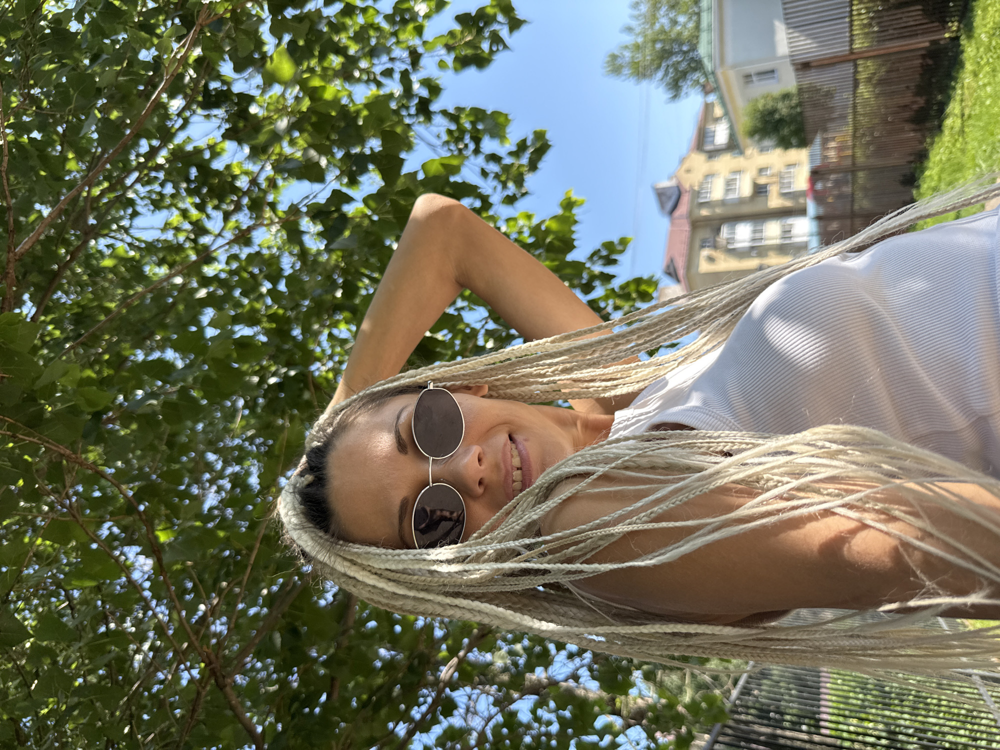 A woman with blonde braided hair and sunglasses is smiling at the camera, with her arm behind her head. She is wearing a white tank top. The background is blurred, showing green leaves from a tree, a blue sky, and buildings in the distance. The scene appears to be outdoors, possibly in a park or yard. The location is identified as Rusanovo, Russia.