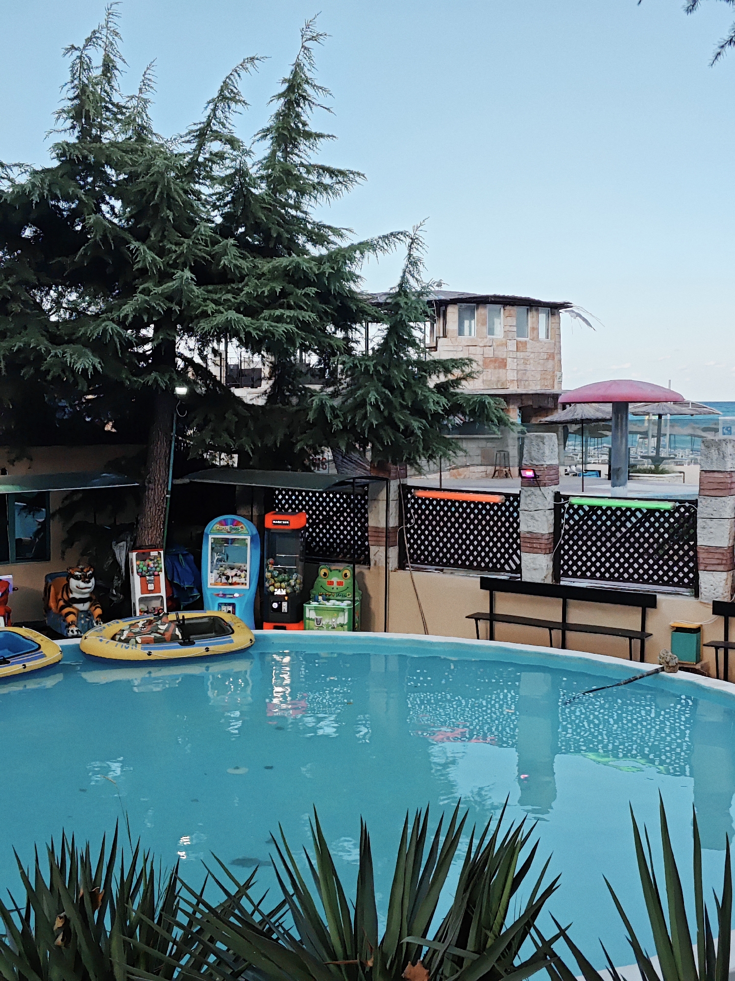 The image shows a bright blue swimming pool with two yellow inflatable boats in it. A large green tree looms over the pool on the left, with its branches reaching towards the sky. Behind the tree, there is a building made of stone with windows. In the background, there are more trees, beach umbrellas, and the ocean can be seen in the distance. Several arcade machines, including a "Super Lucky" claw machine and a "Tiger" themed machine, are lined up near the edge of the pool. The overall scene suggests an amusement area or resort, possibly located near a beach. The sky is clear and pale blue, and the lighting suggests it is either early morning or late evening.