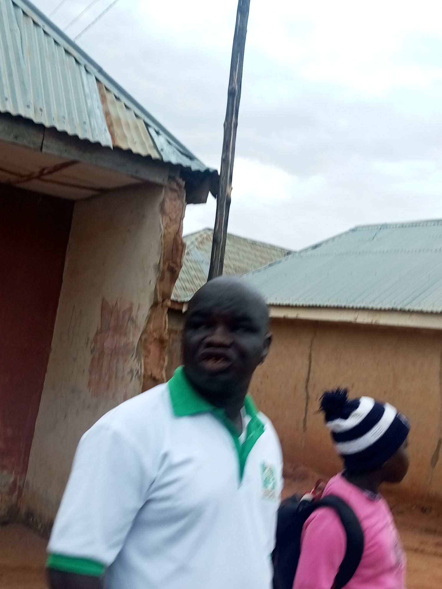 A medium shot of a Black man and a child walking in an outdoor setting in Gombe, Nigeria. The man is in the foreground, partially blurred, wearing a white polo shirt with green trim on the collar and sleeves. A small logo is visible on the left side of the shirt. The child is slightly behind and to the right of the man, wearing a pink shirt, a black backpack, and a blue and white striped beanie with a pom-pom.
In the background are buildings with corrugated metal roofs. The building on the left has a wall with damaged plaster showing exposed brick underneath. A utility pole stands tall, bisecting the sky. The sky is overcast and grey. The ground appears to be dirt or reddish soil.