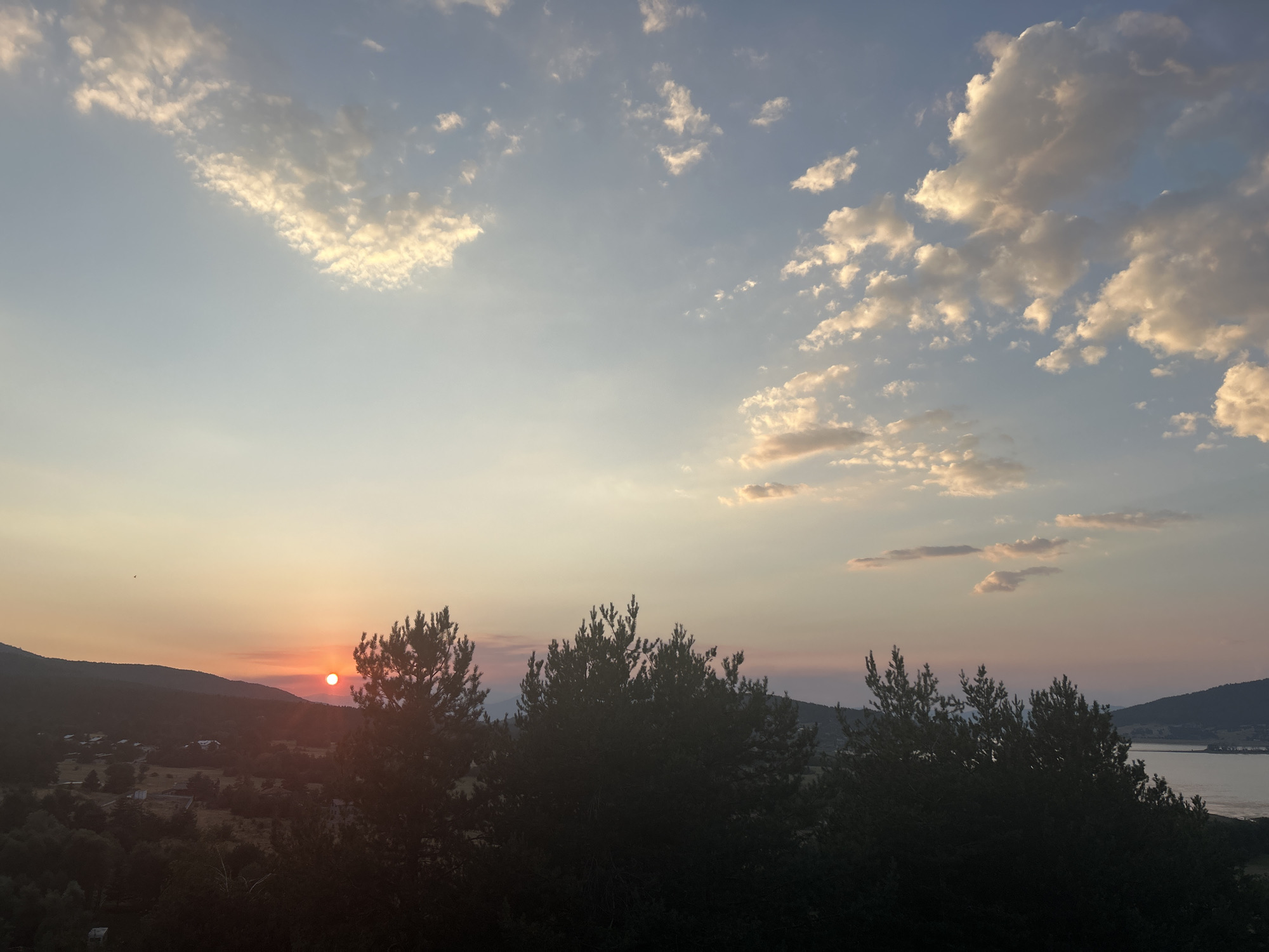 The image depicts a sunset over Batak, Bulgaria. In the foreground, silhouetted pine trees dominate the view, with a few buildings and sparse vegetation visible in the middle ground. The sun, a bright orange orb, is partially obscured by the horizon and a thin layer of haze. In the distance, rolling hills form the backdrop, and a body of water, likely Batak Reservoir, is visible on the right. The sky is a gradient of pale blue and orange, with scattered clouds catching the last rays of the sun. The overall impression is one of serene natural beauty.