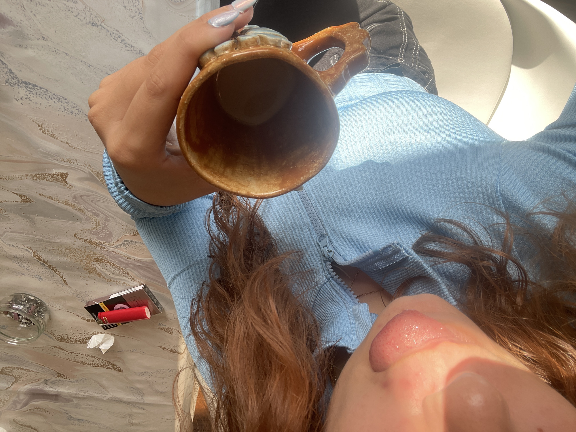 A photograph taken from an elevated perspective depicts a person seated at a table, holding a ceramic mug. The person is wearing a light blue, ribbed top and dark-colored pants. The table surface is a swirling pattern of white, beige, and light brown. On the table, near the edge, is a small glass jar containing small objects, a packet of cigarettes, a red lighter, and a crumpled piece of paper. The person's face is partially visible, showing the nose and upper lip. The person's hair is long and reddish-brown. The background is blurred, showing a white chair. The location in the city of Saarbrücken, Germany, cannot be determined from the image.