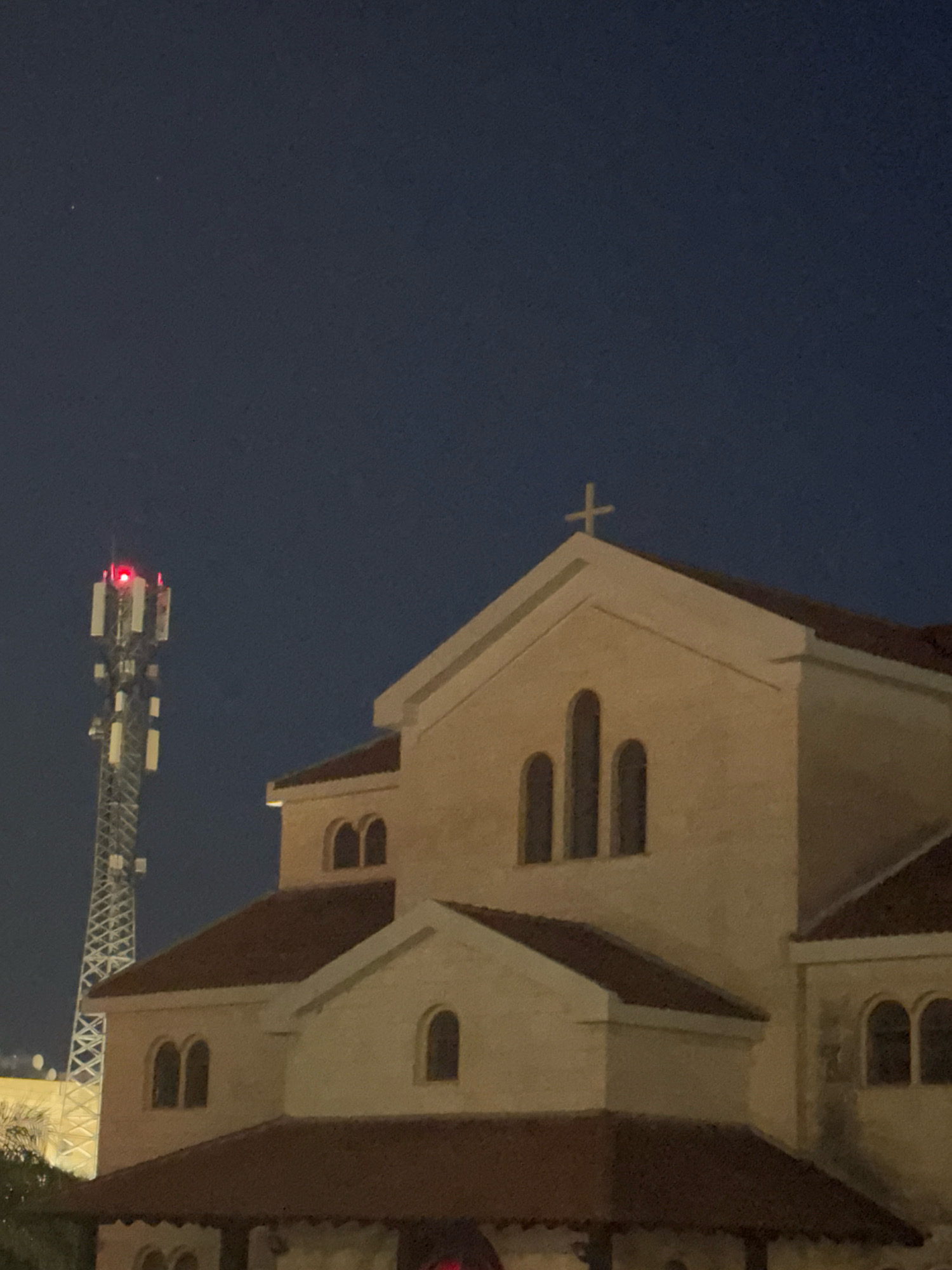 The image depicts a cellular tower with red flashing lights on top, positioned to the left of a light-colored building that appears to be a church, evidenced by a cross on its roof. The building has arched windows and a reddish-brown tiled roof. The sky is dark, suggesting it is nighttime. The scene is set in Germasogeia Municipality, Cyprus.